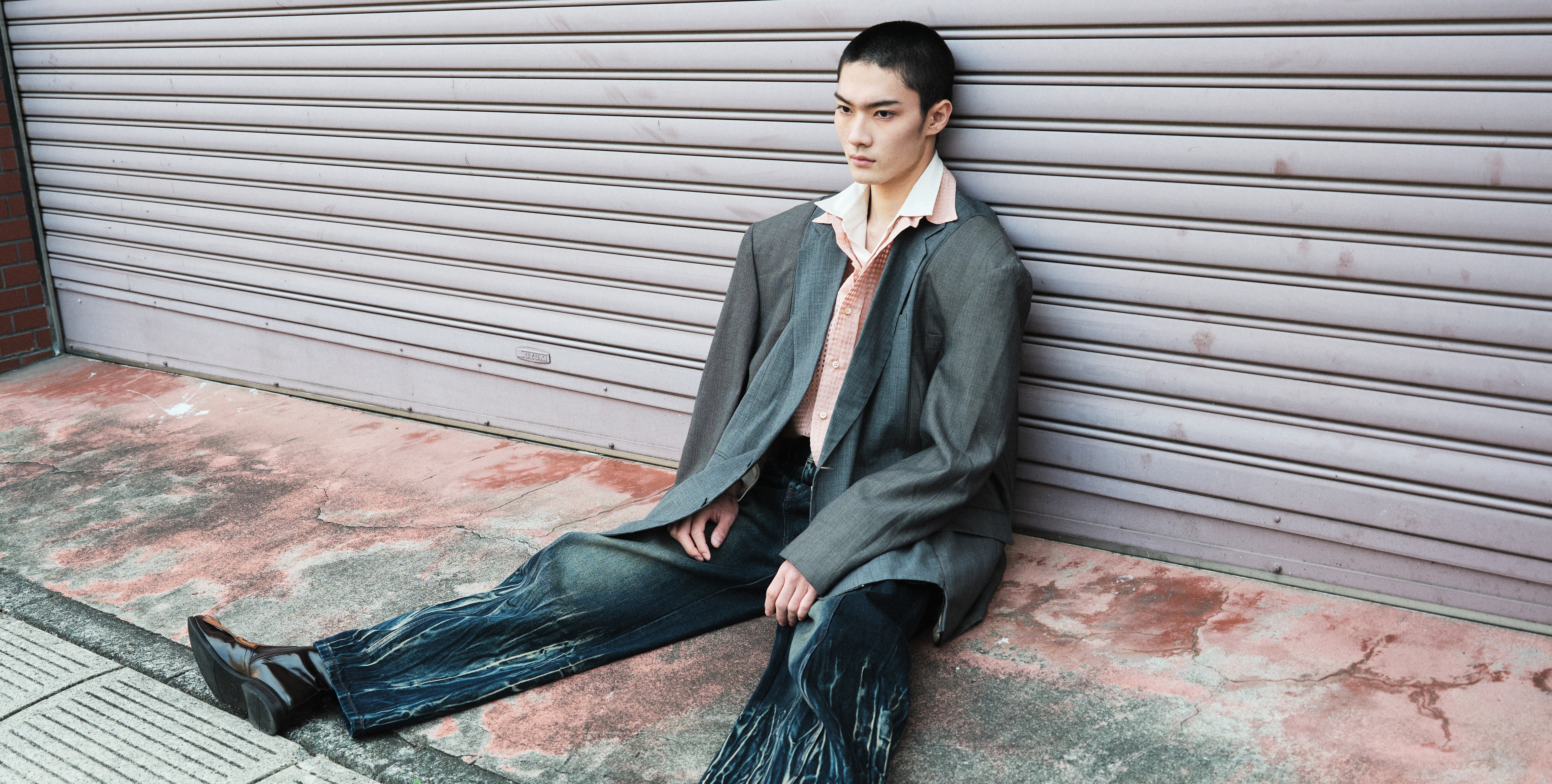 Person in grunge style outfit sits against a garage door, urban fashion mood.