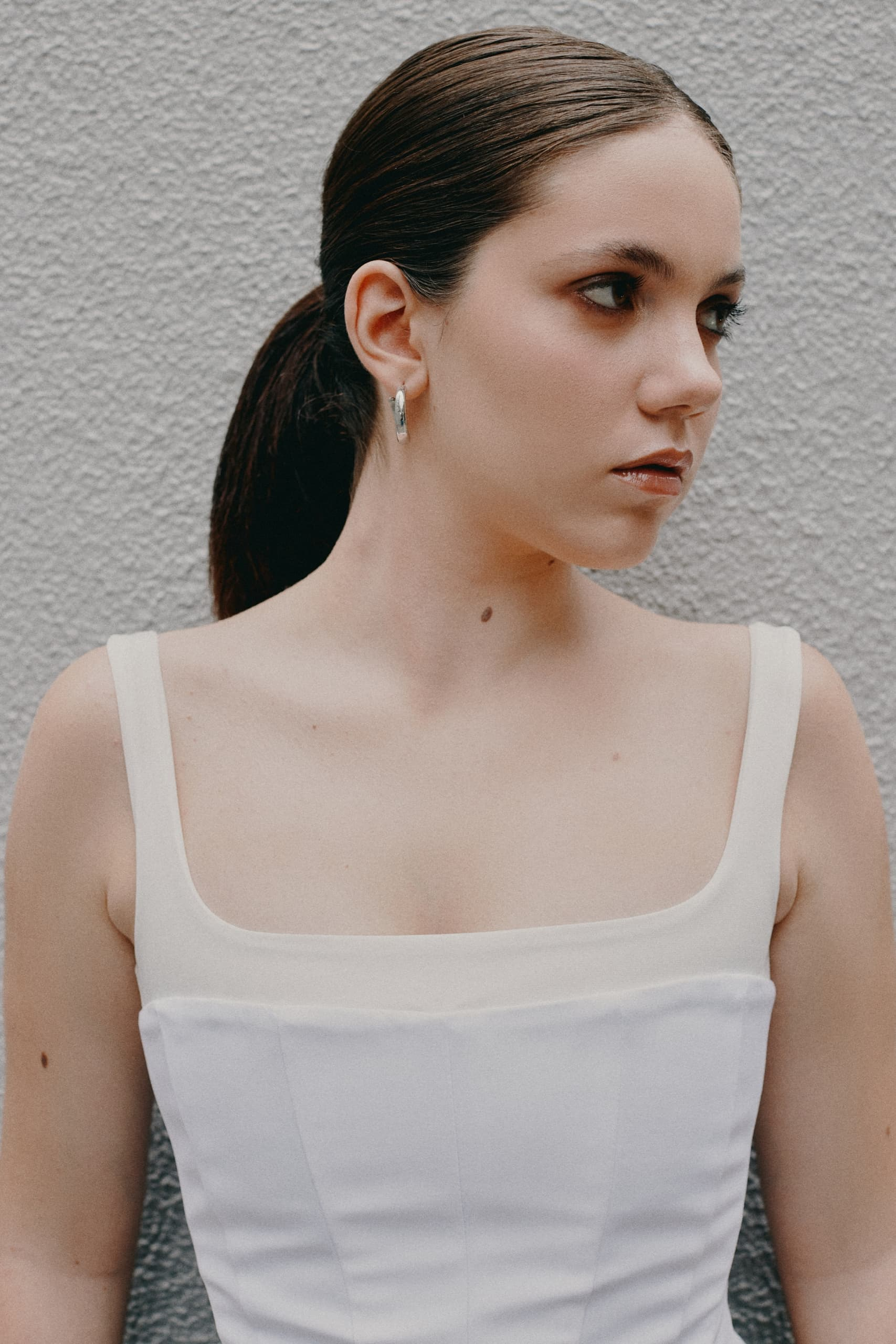 Woman wearing white dress, looking left against textured wall.
