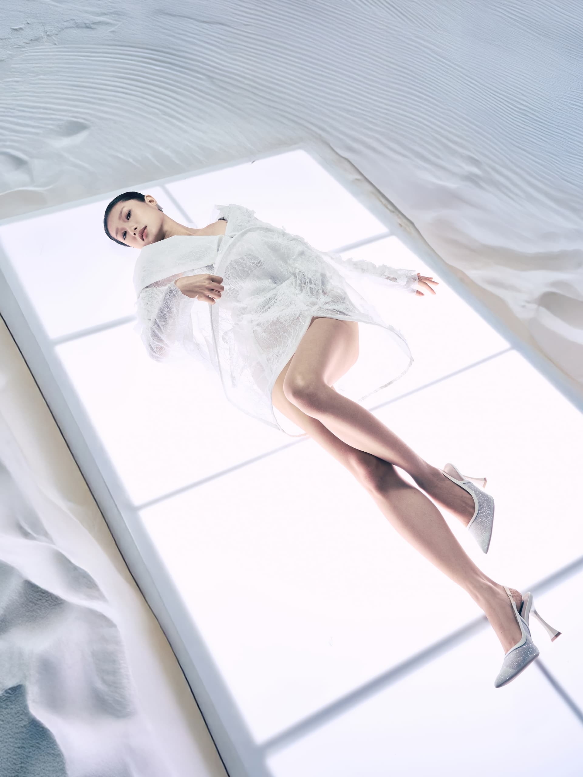 Woman in white lace dress and heels, lying on backlit glass floor.