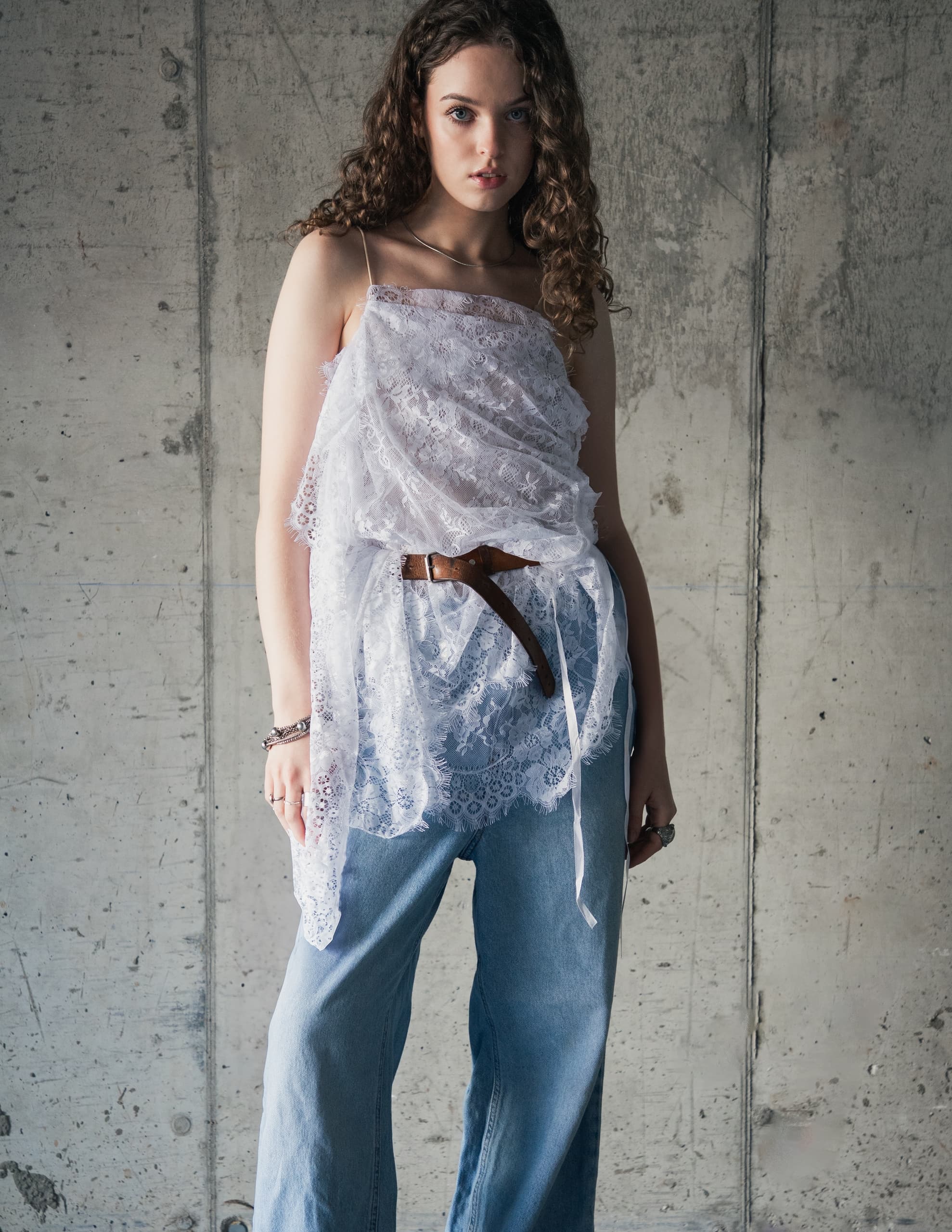 Woman in lace top and jeans standing against concrete wall.