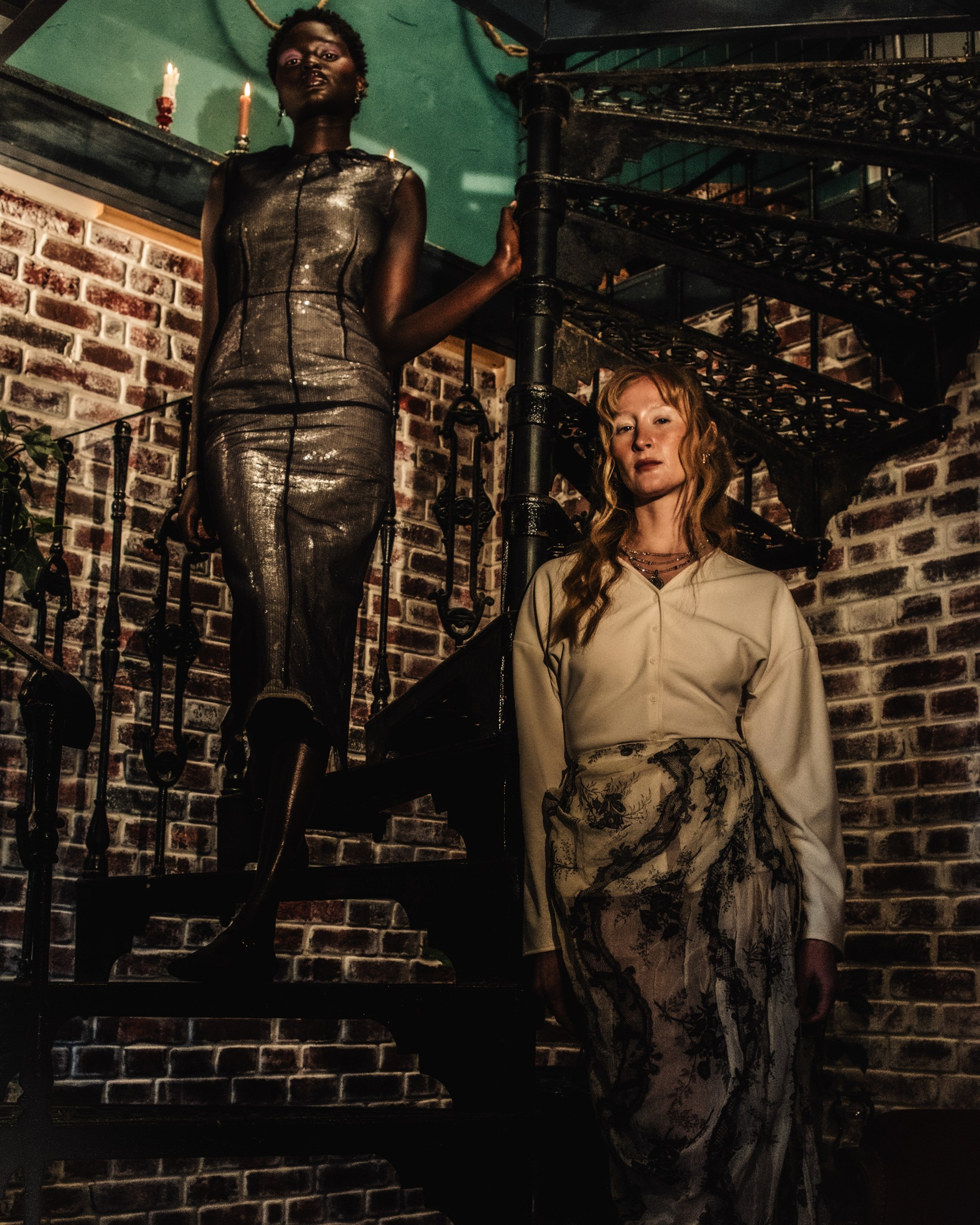 Two models on spiral staircase, elegant fashion, brick wall background.