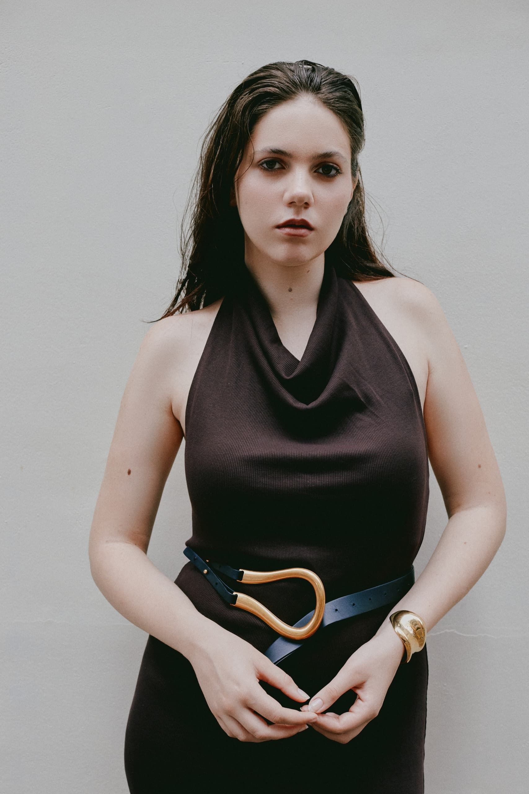 Woman in a black dress with a unique belt and gold bracelet against a plain background.