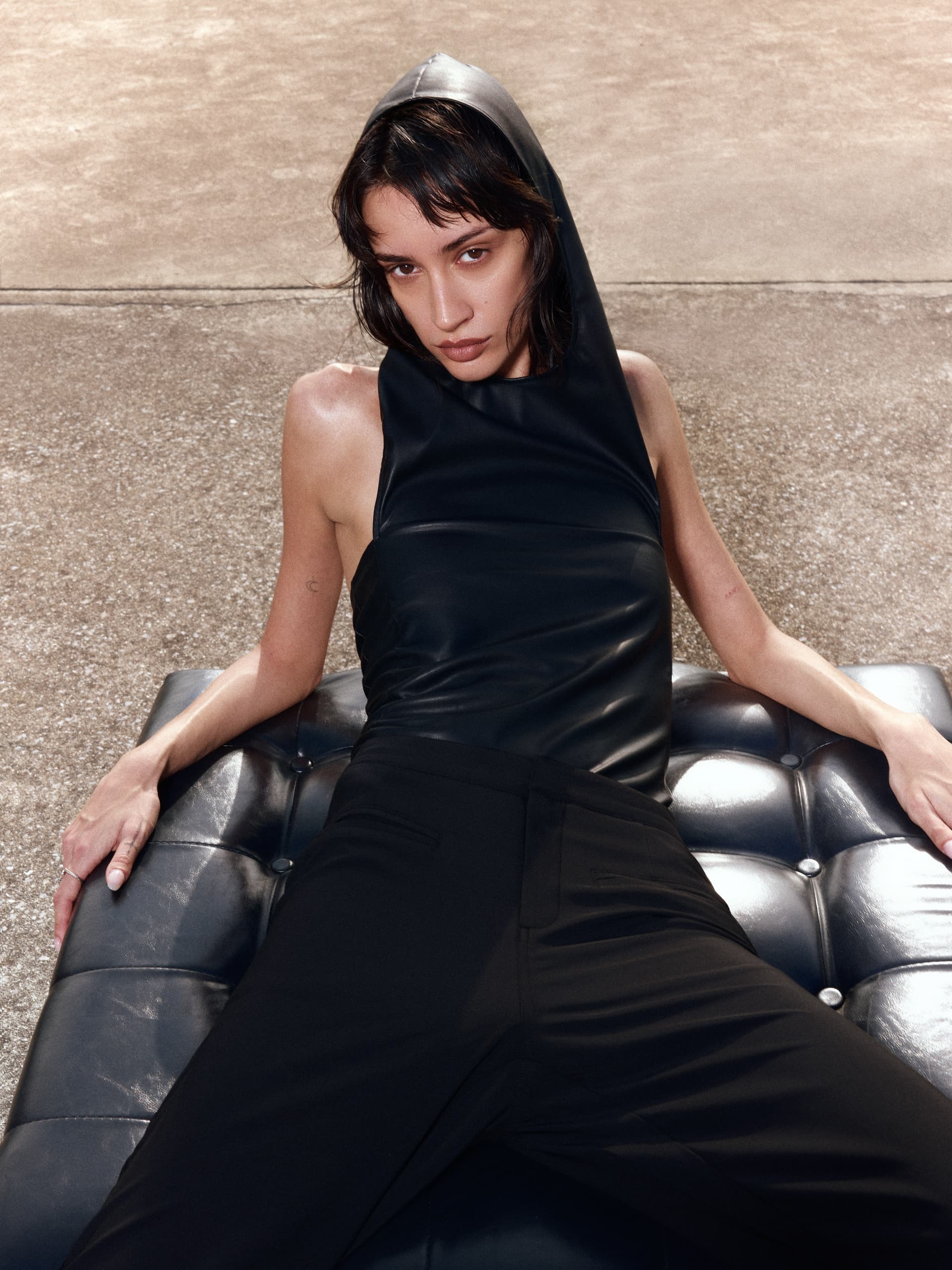 Model in black leather hooded top reclines on a tufted bench outdoors.