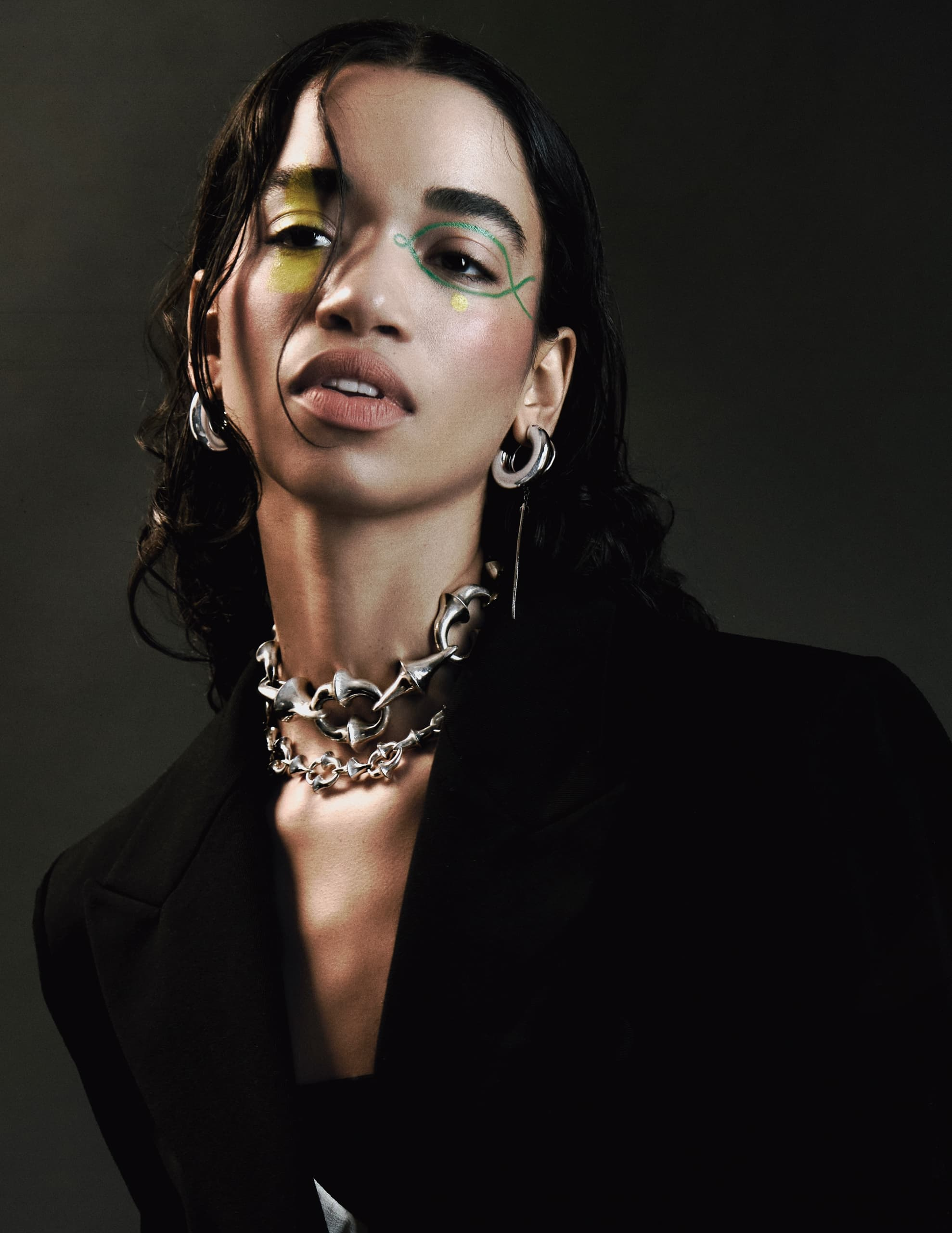 Person in dark attire with bold makeup and silver jewelry in a studio portrait.