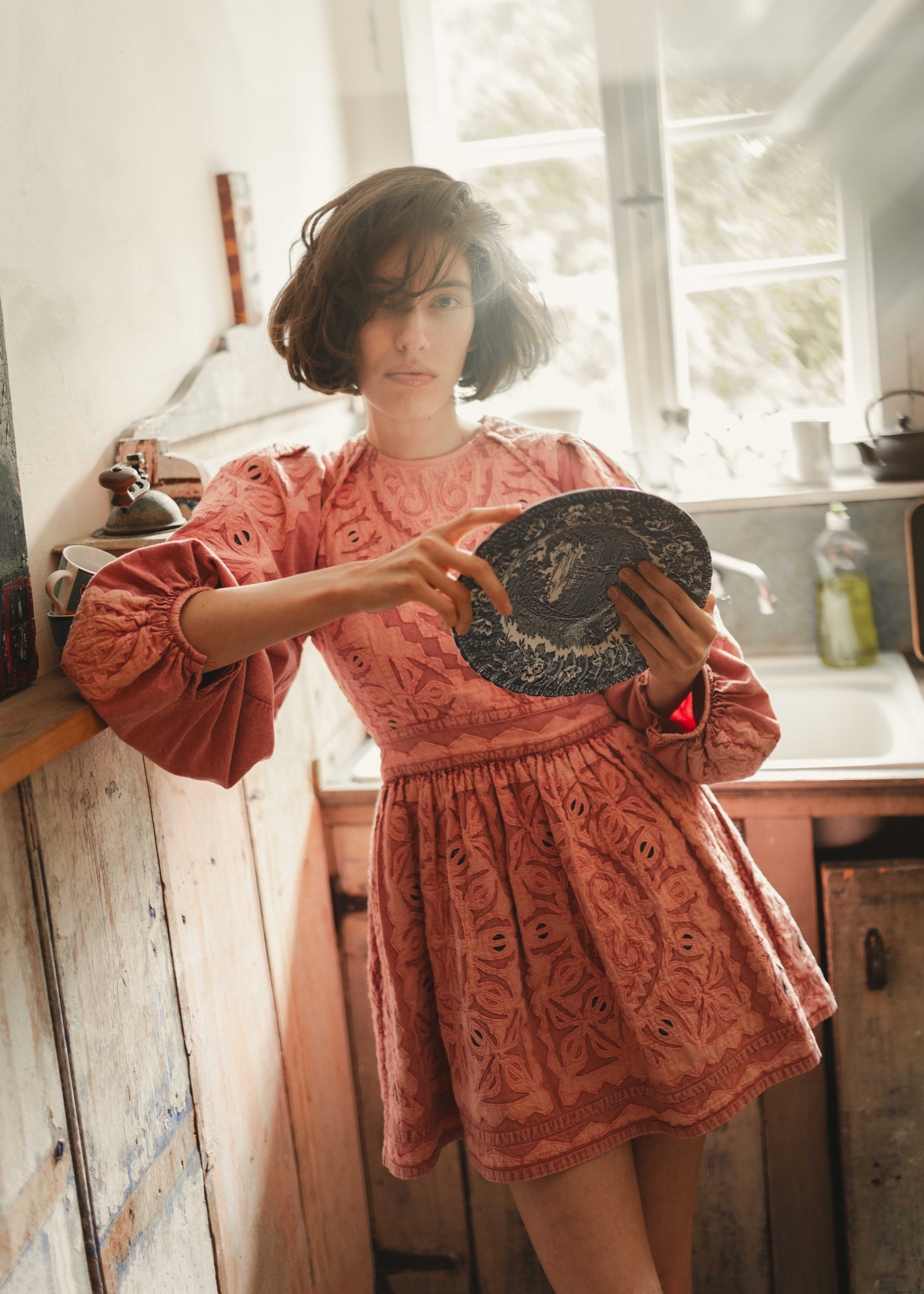 Woman in pink dress holding decorative plate in rustic kitchen setting.