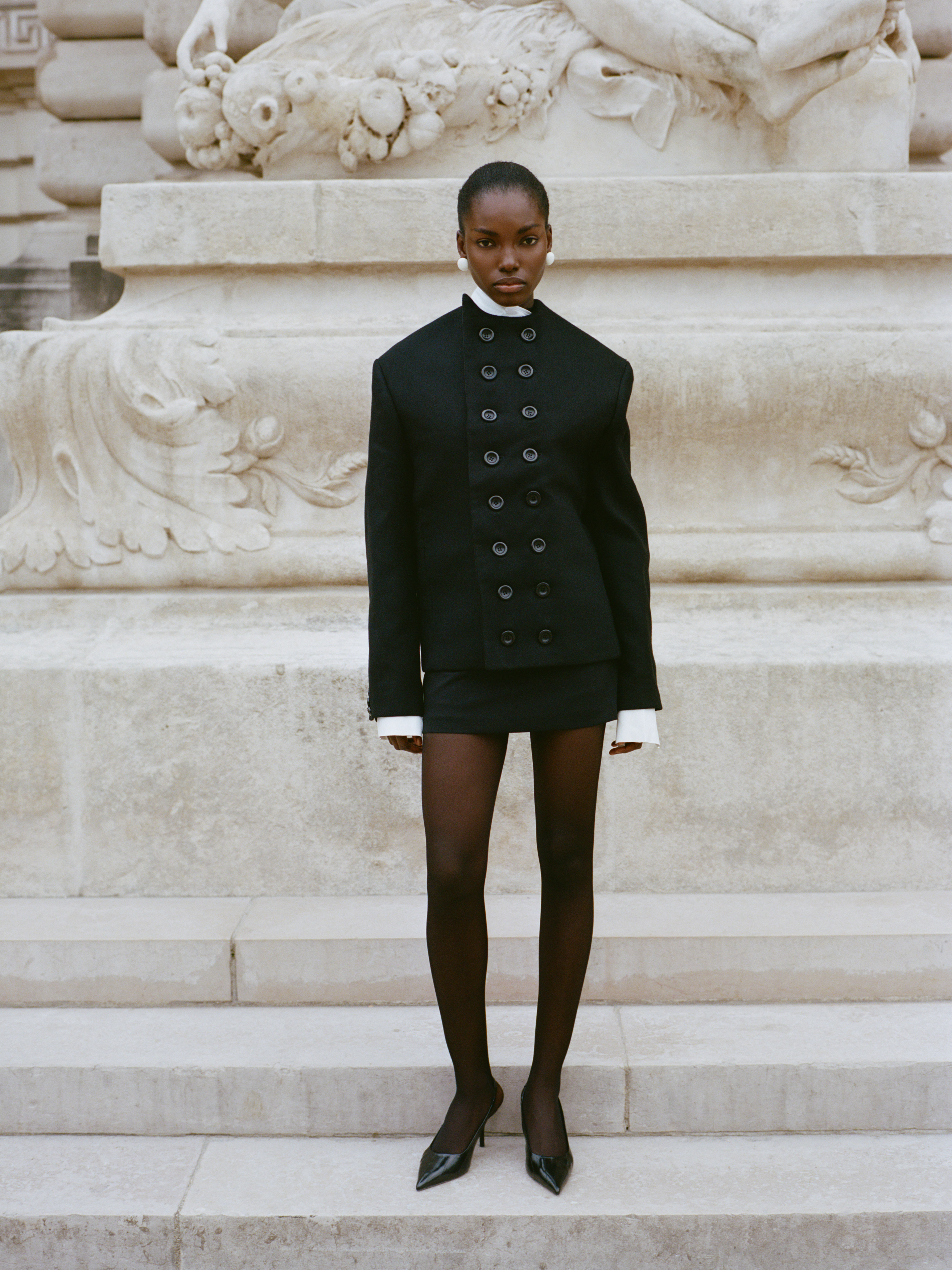 Model in stylish black outfit stands before ornate stone sculpture.