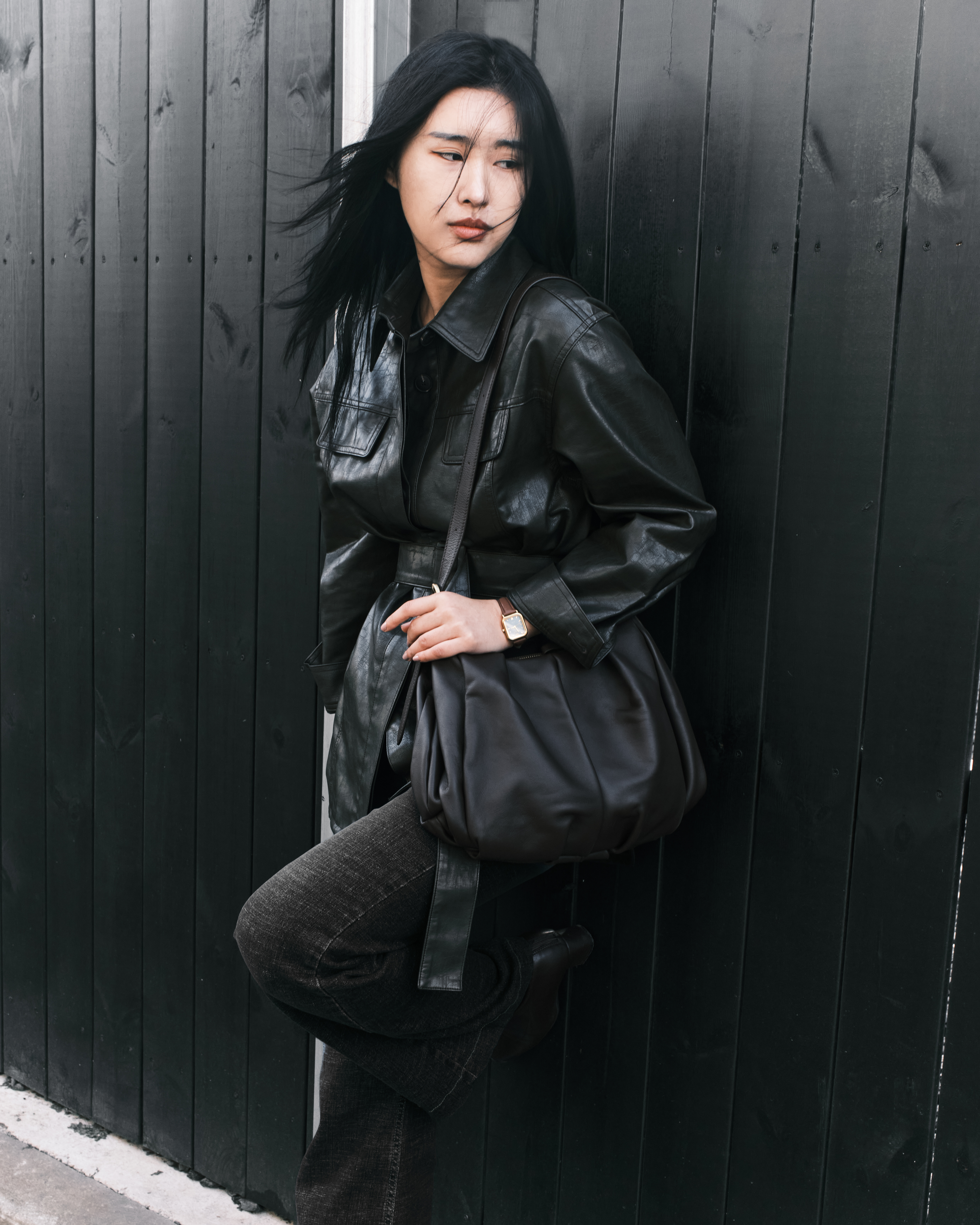 Woman in black leather jacket leans on a dark fence, holding a stylish bag.