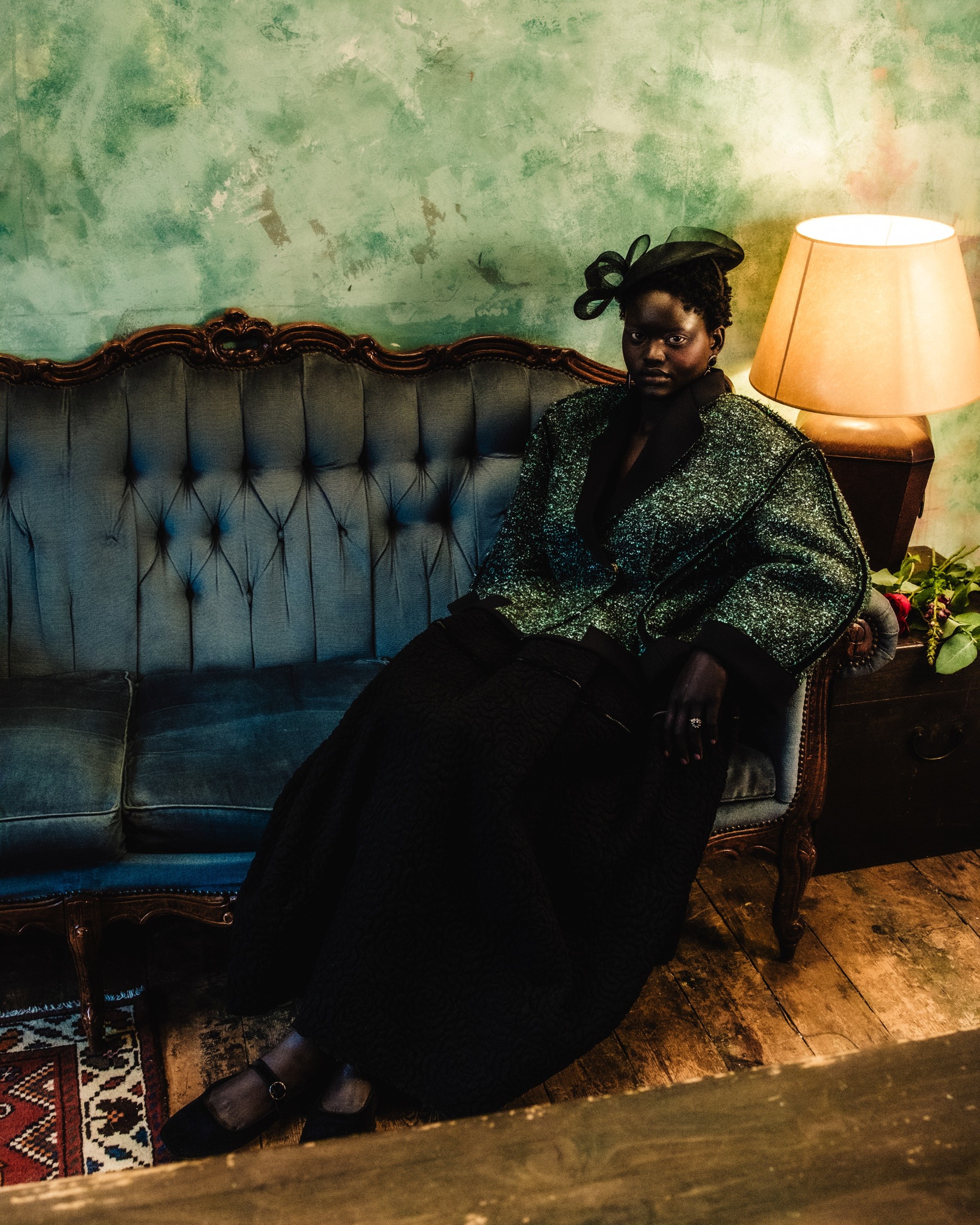 Person in elegant outfit sitting on vintage blue sofa near a lamp.