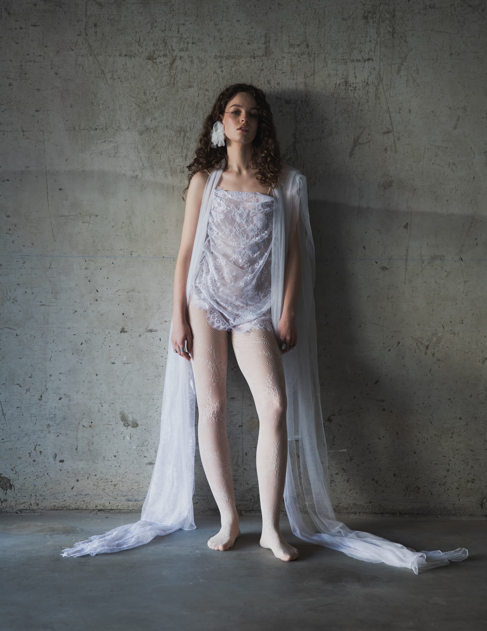 Model in sheer lace outfit against textured concrete wall. Fashion photoshoot.