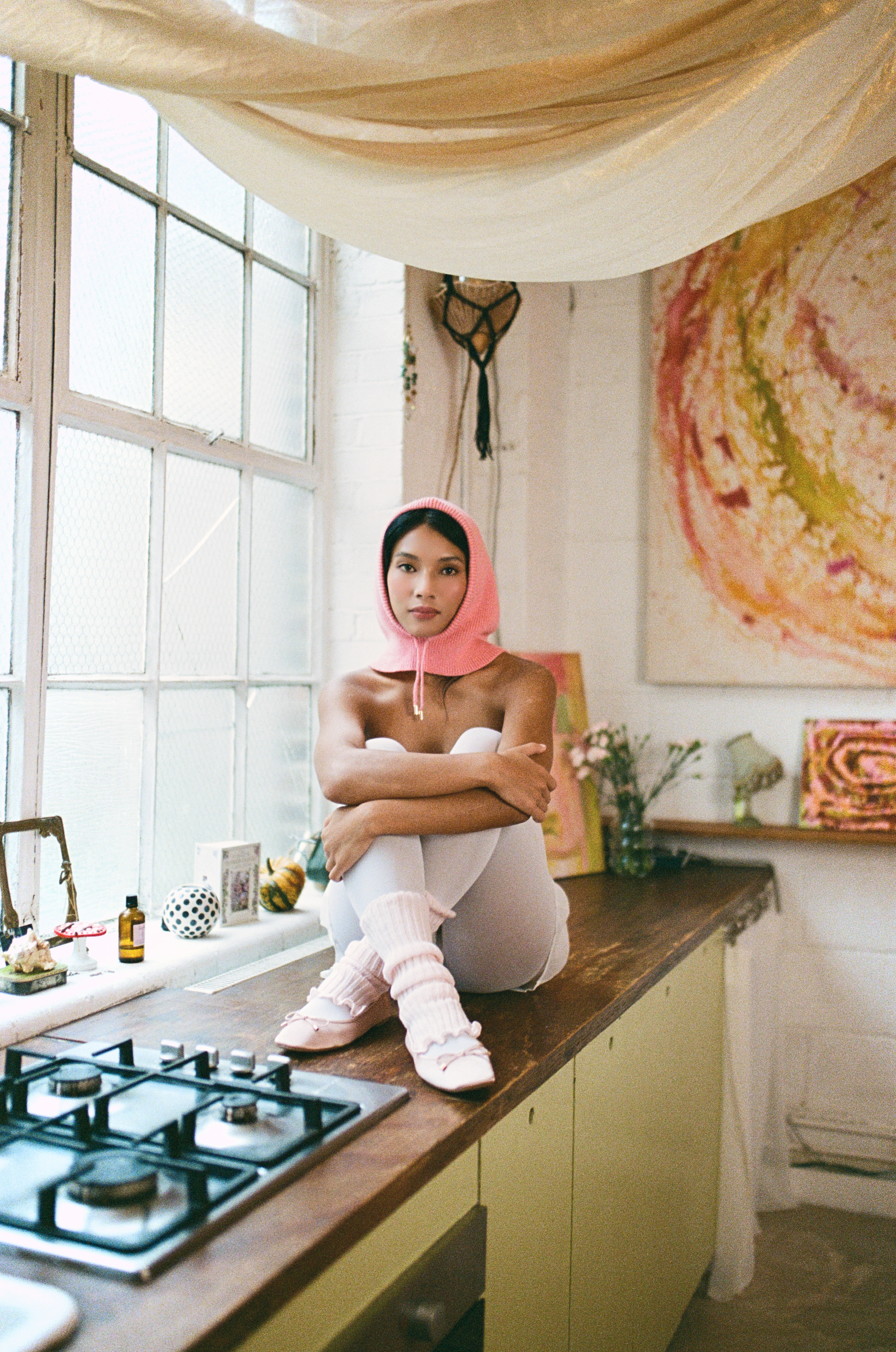 Person in pink hood sitting on counter in artsy kitchen setting.