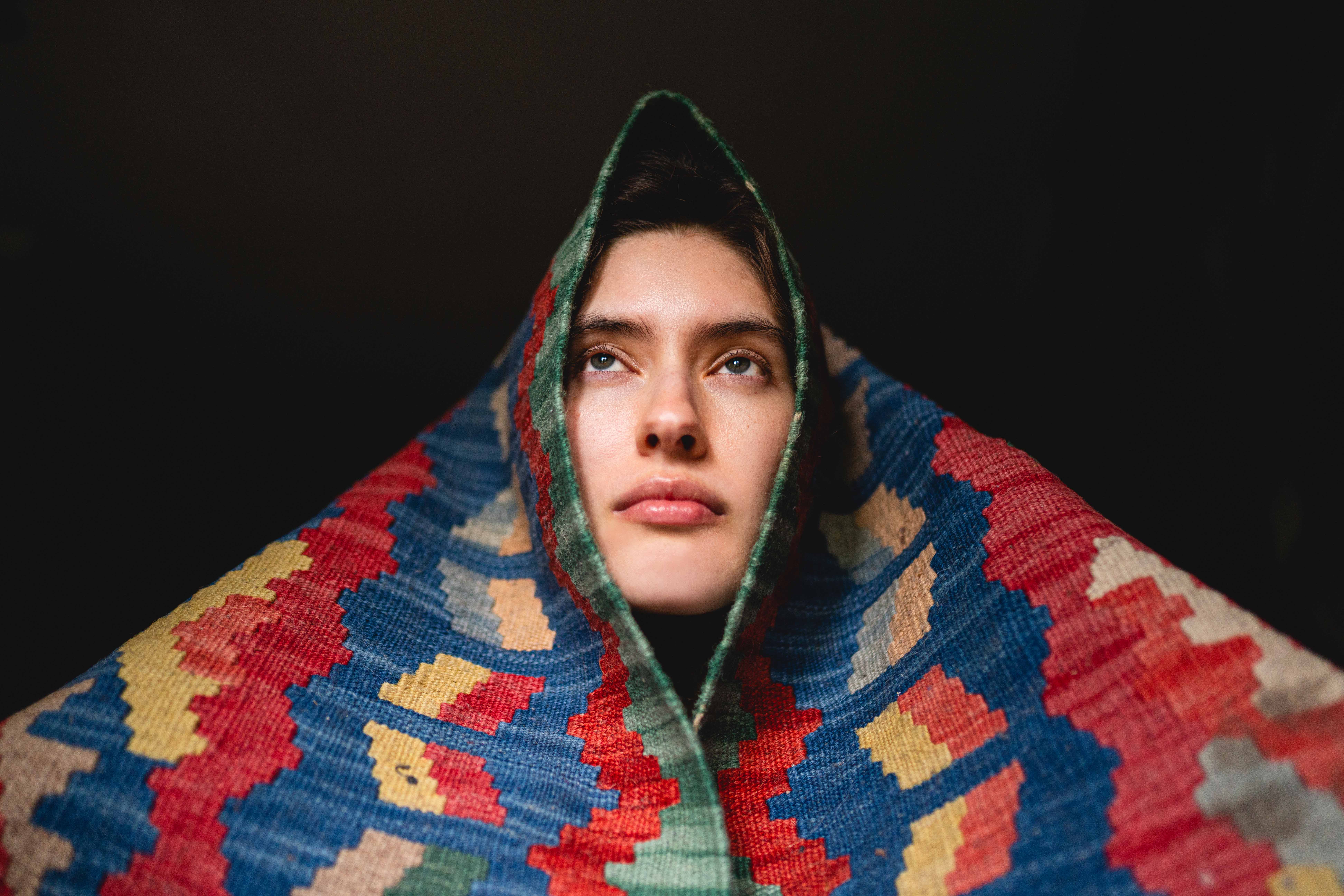 Person wrapped in colorful patterned fabric, gazing upward against a dark background.