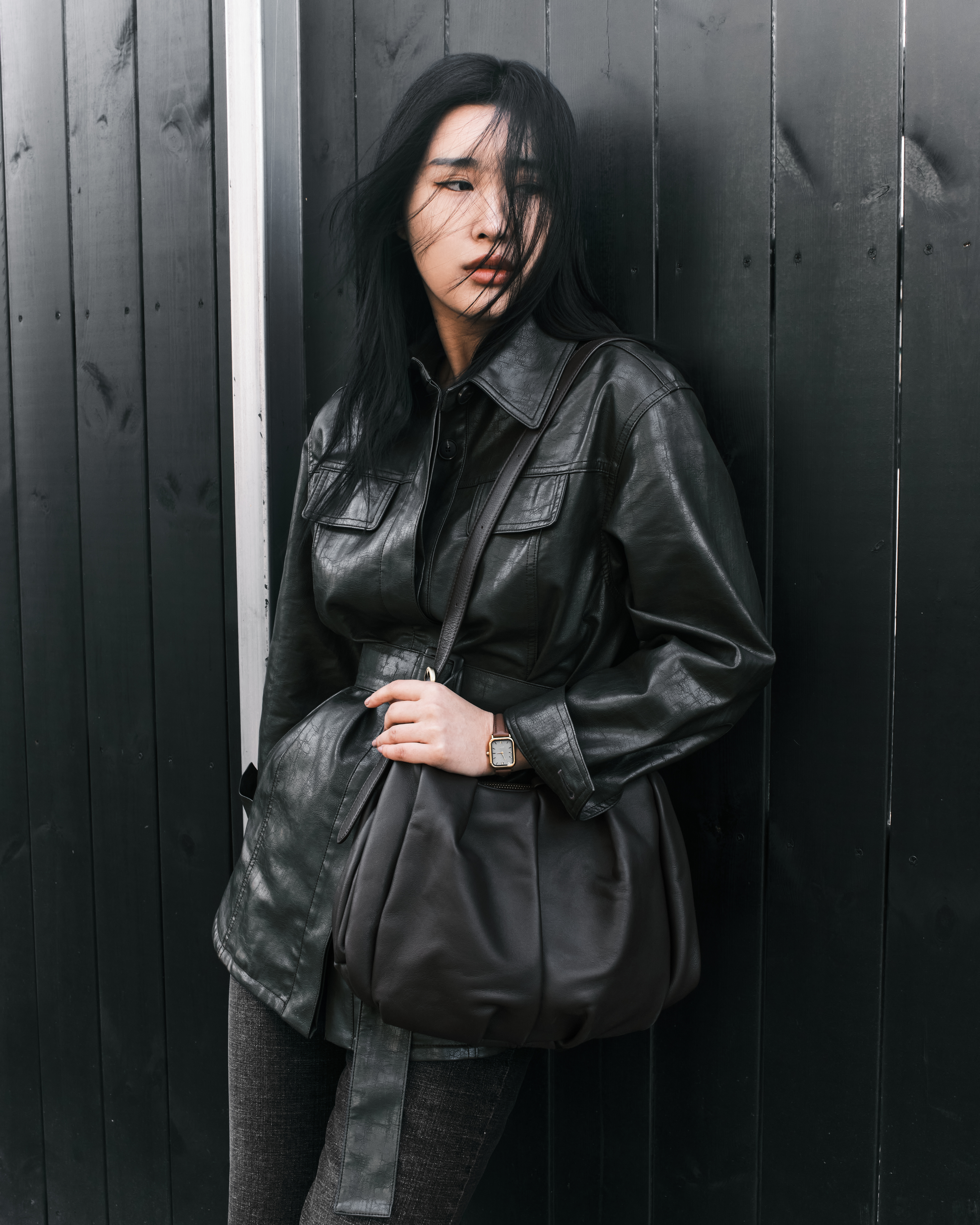 Stylish woman in black leather jacket leaning on dark wooden fence.