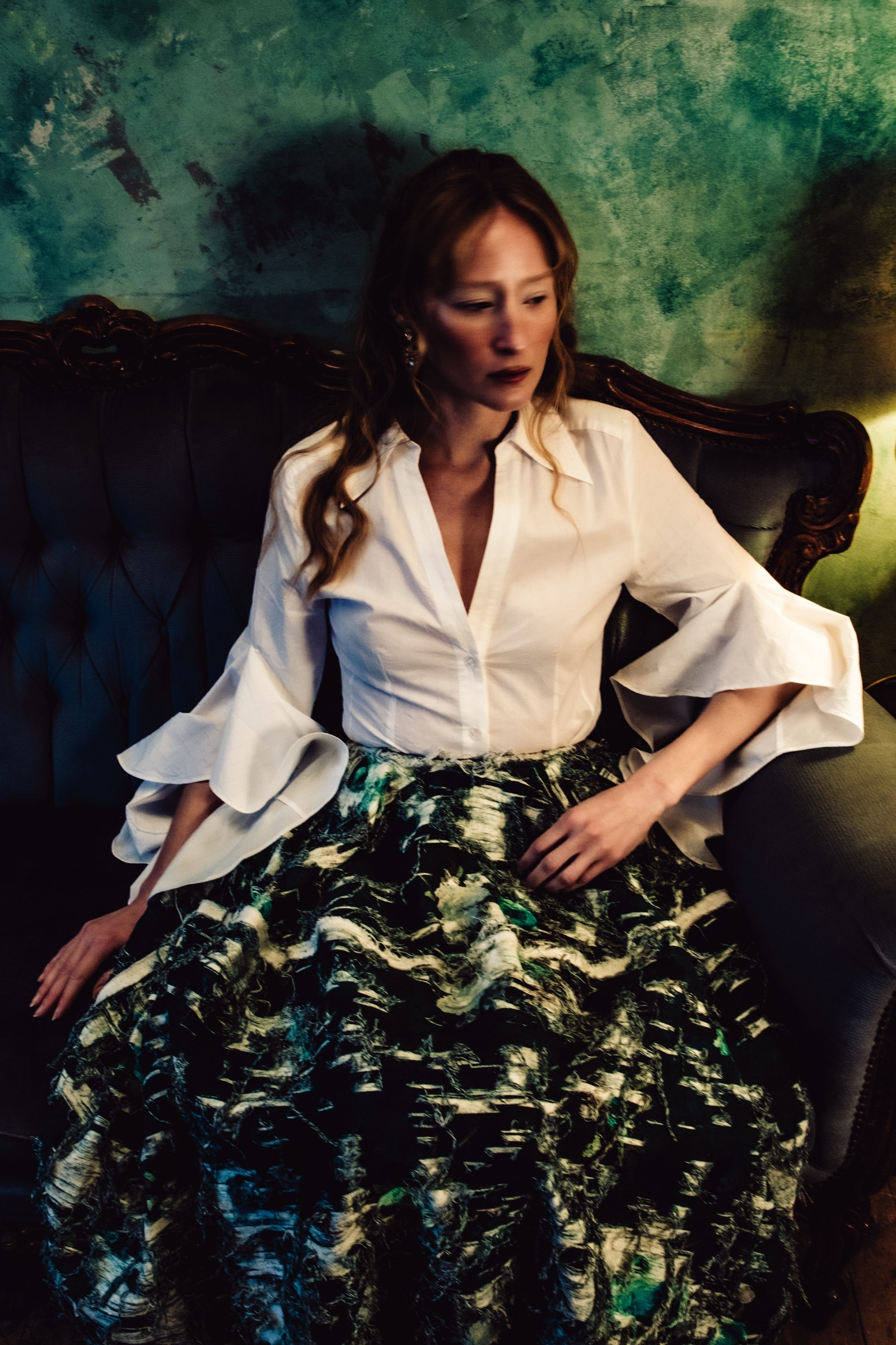 Person in elegant attire sitting on a vintage sofa against a textured green wall.