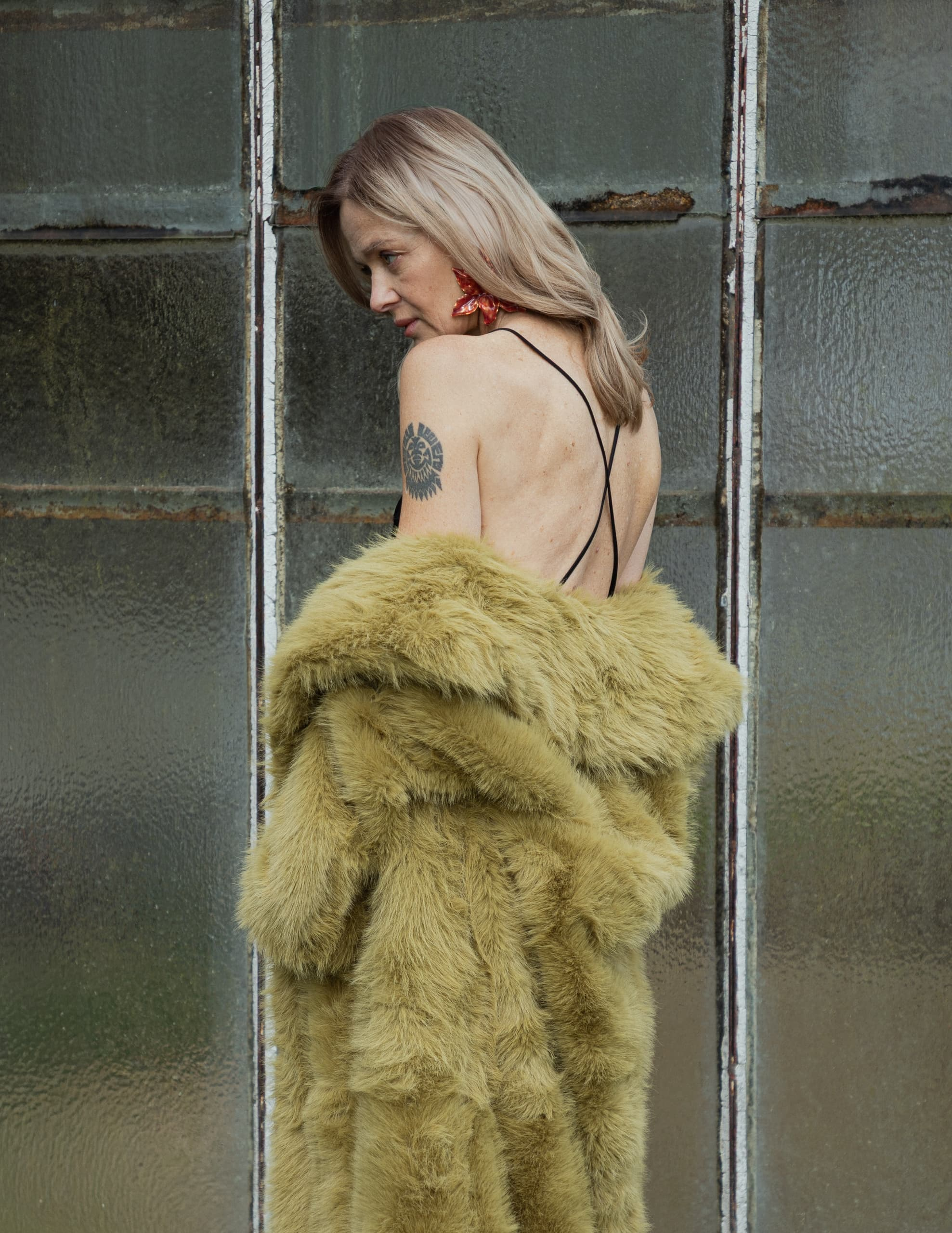 Woman in a backless dress and fur coat stands by a rustic glass door.