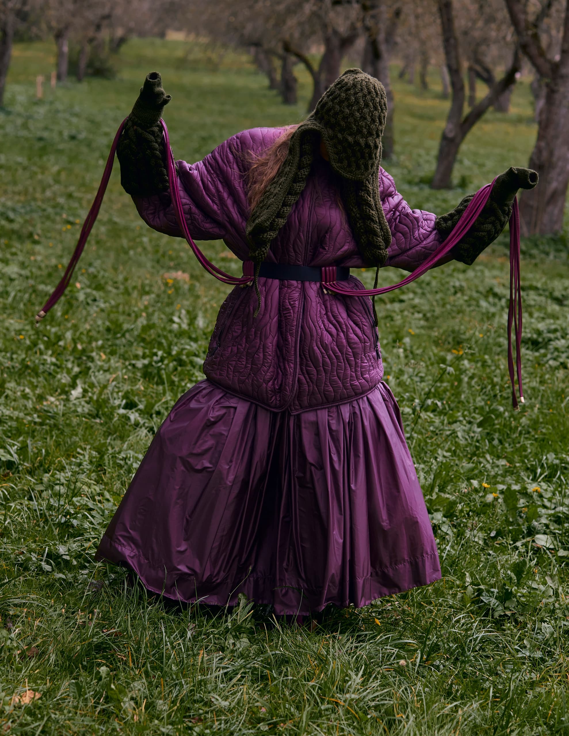 Person in purple quilted outfit and green knit hat in a grassy orchard.