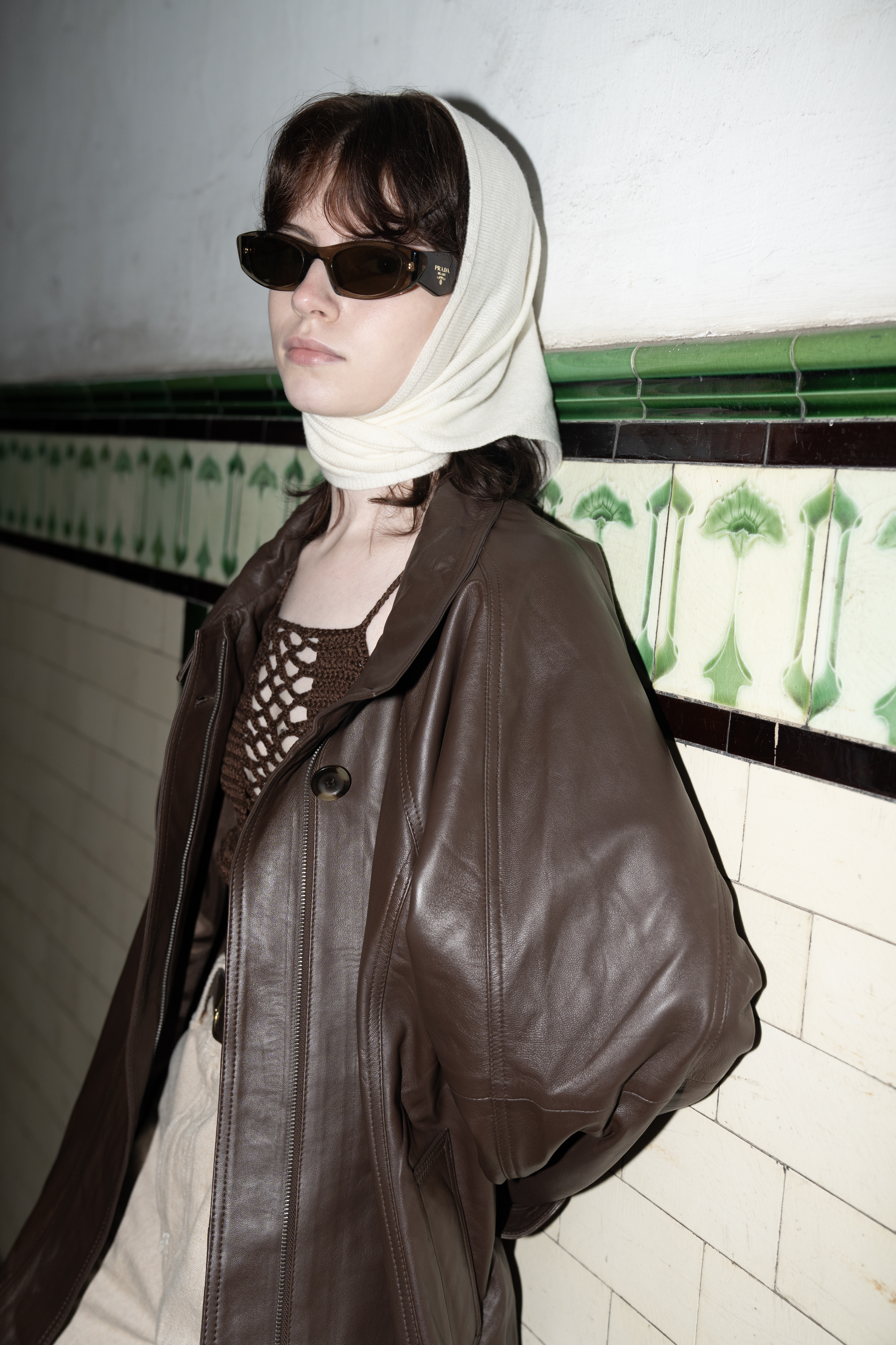 Fashionable woman in a brown leather coat and sunglasses by a tiled wall.