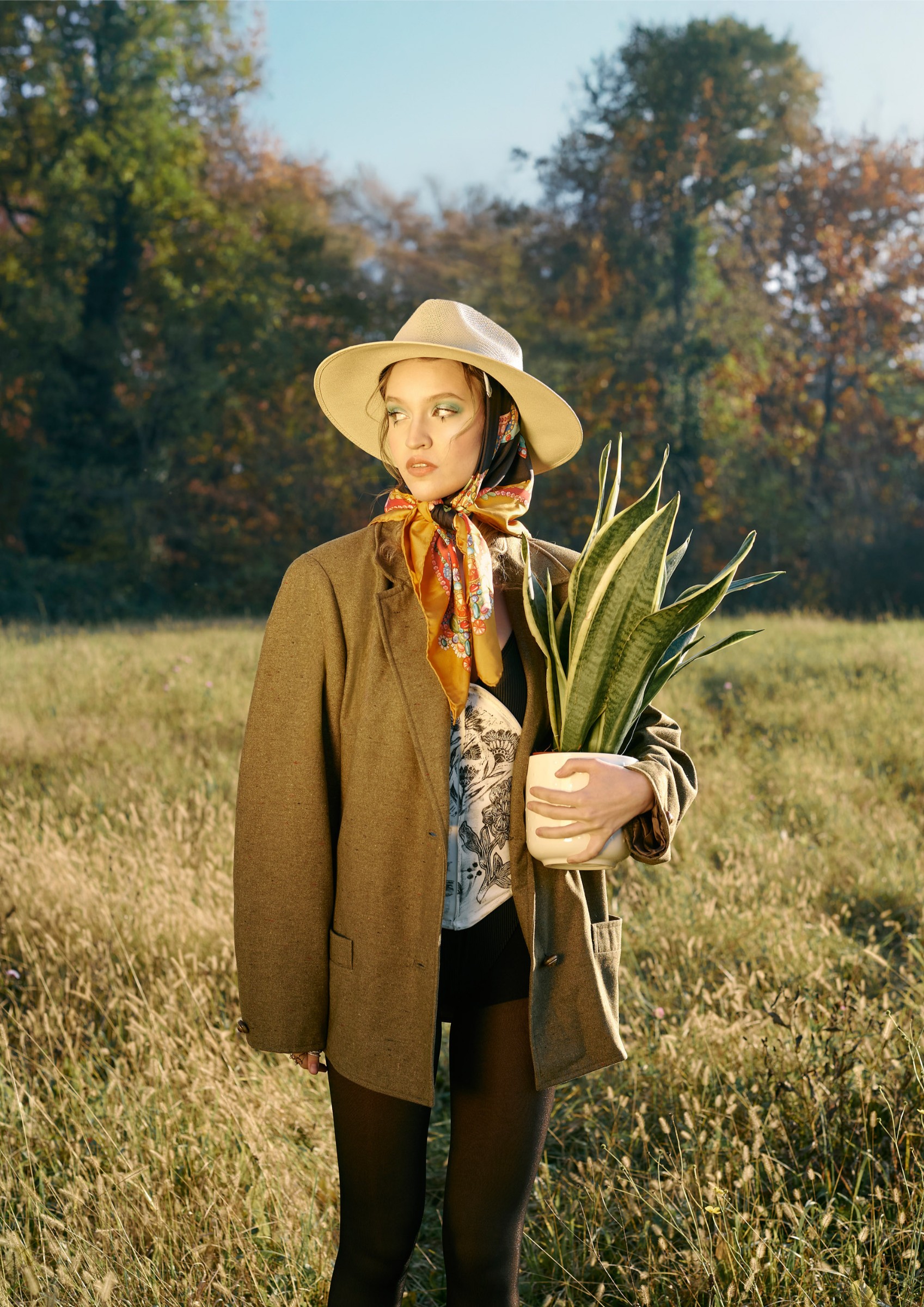 Woman in hat and scarf holds potted plant in sunny field.
