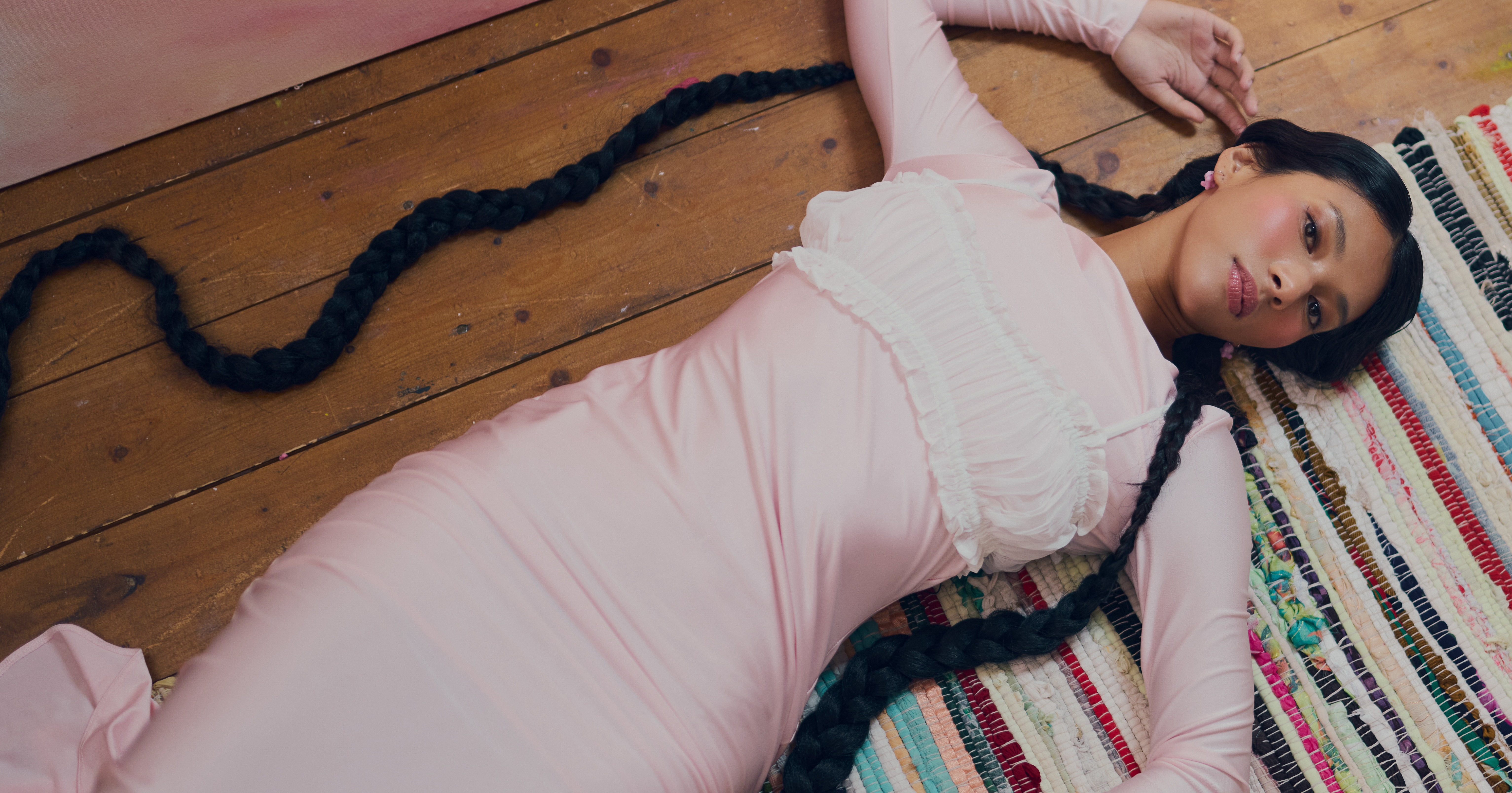 Woman in pink dress lying on a colorful rug, long braids spread on wooden floor.