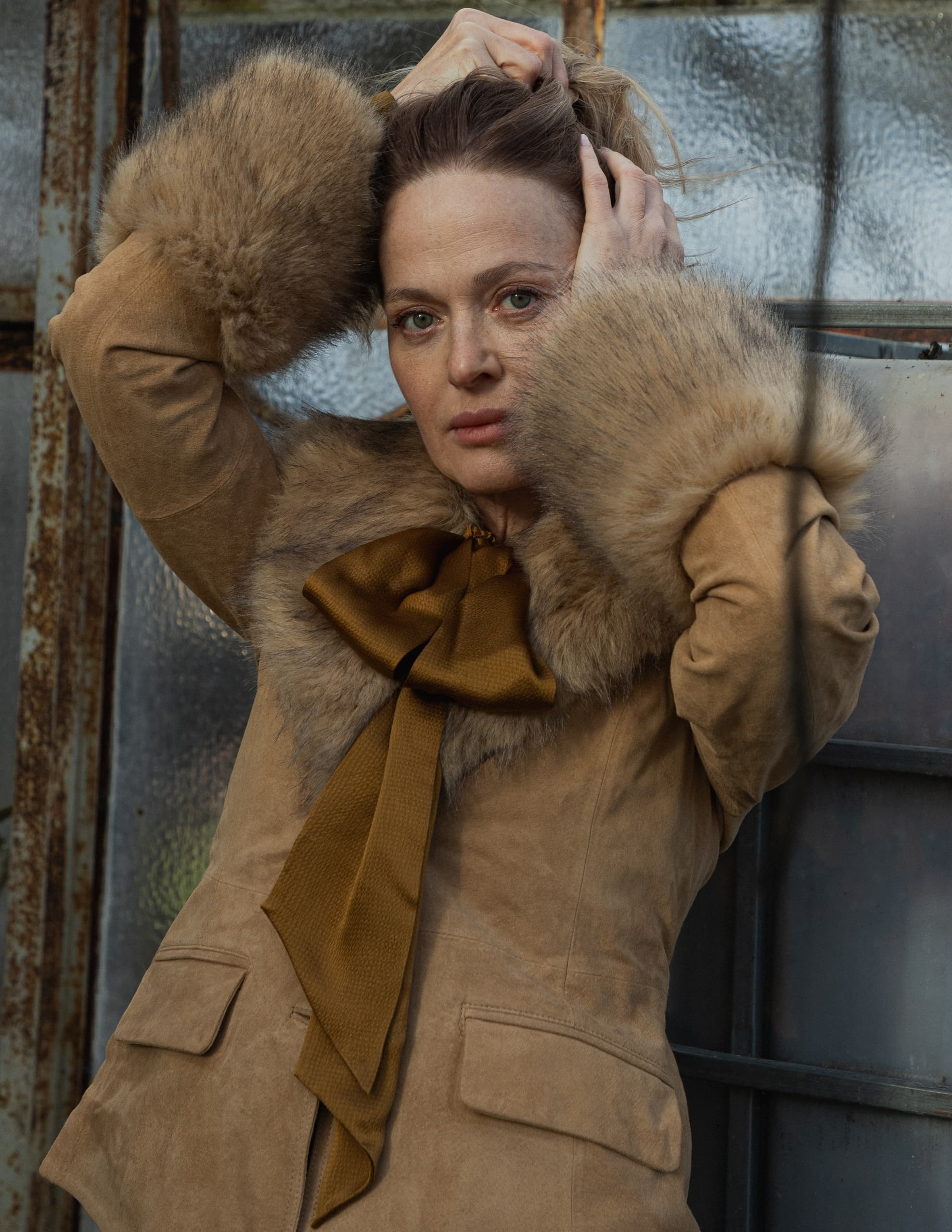 Woman in fur-trimmed coat poses against rustic window background.