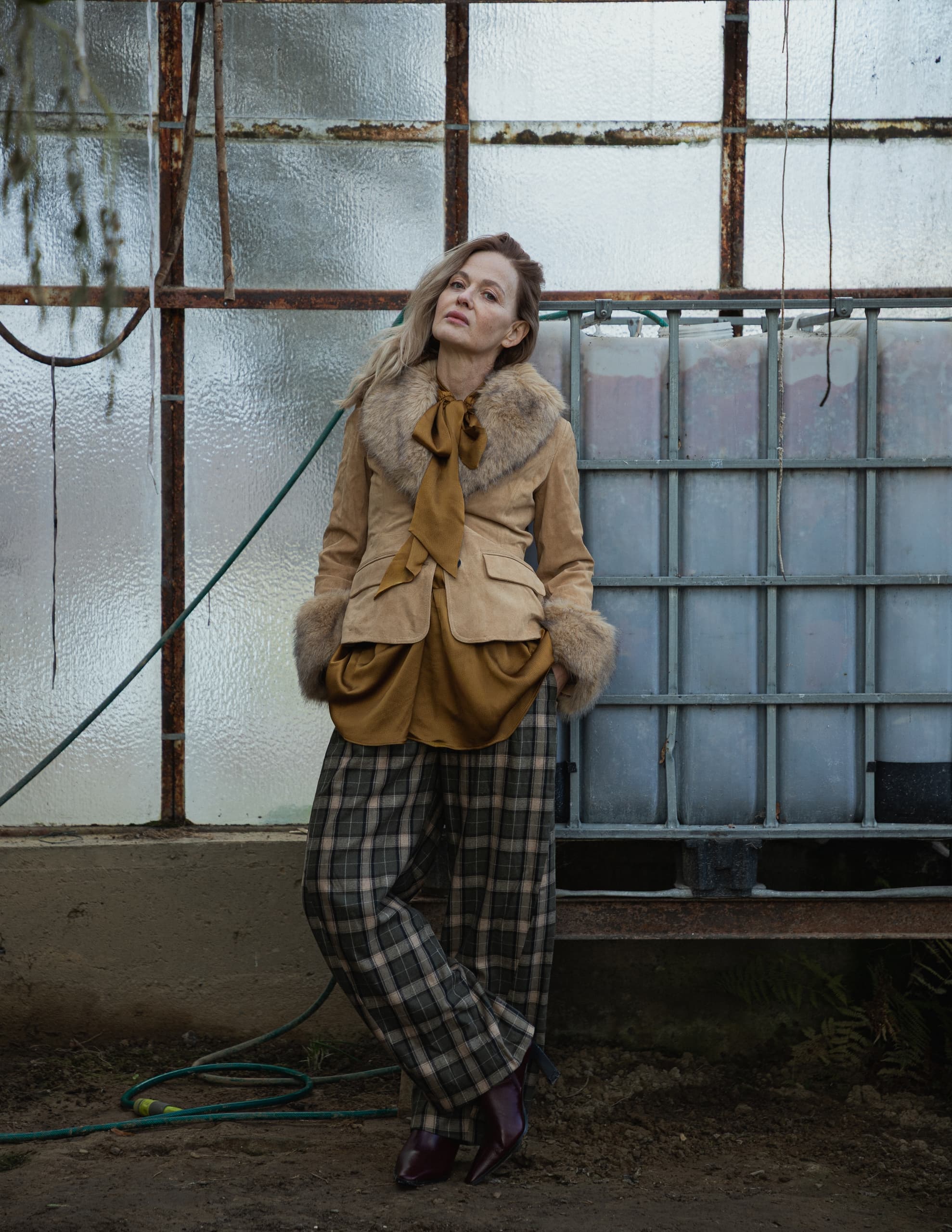 Person in stylish fur coat and plaid pants leaning in rustic industrial setting.