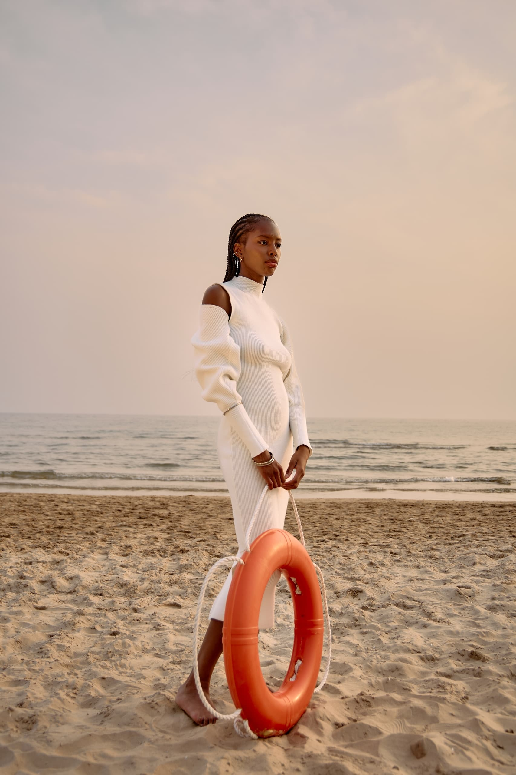Woman in white dress with lifebuoy on sandy beach at sunset.