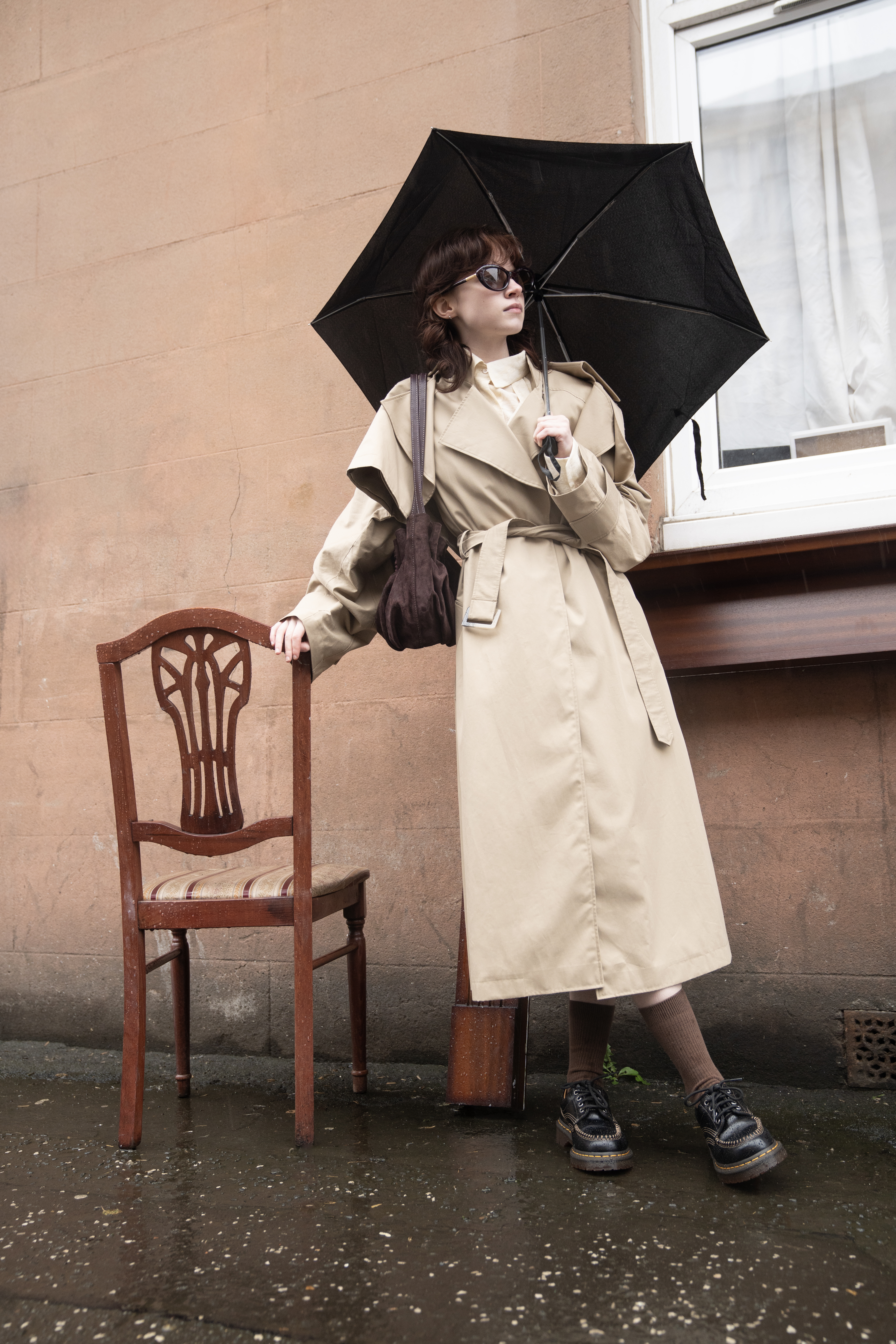 Person in trench coat holding umbrella by chair on rainy street.