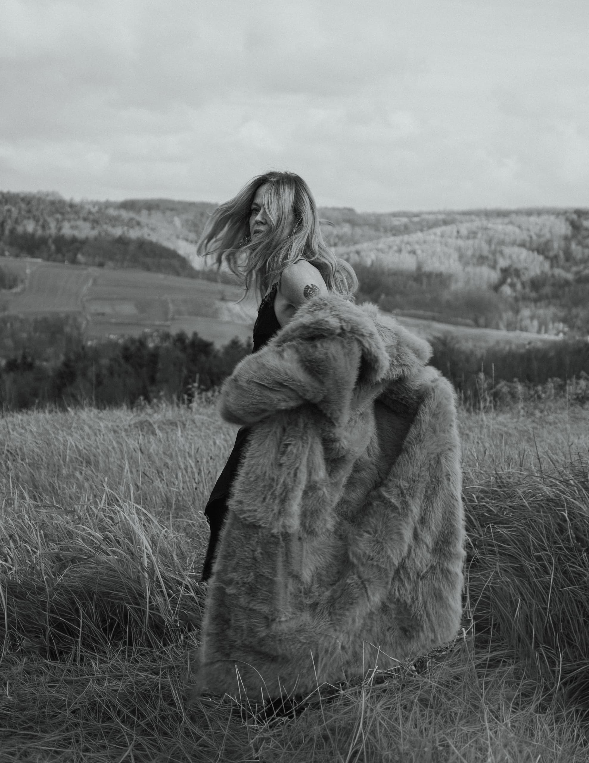 Woman in fur coat standing in windy field, black and white landscape scene.