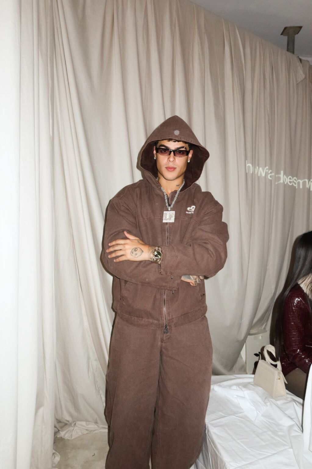 Man in a brown hooded outfit and sunglasses, standing with arms crossed.