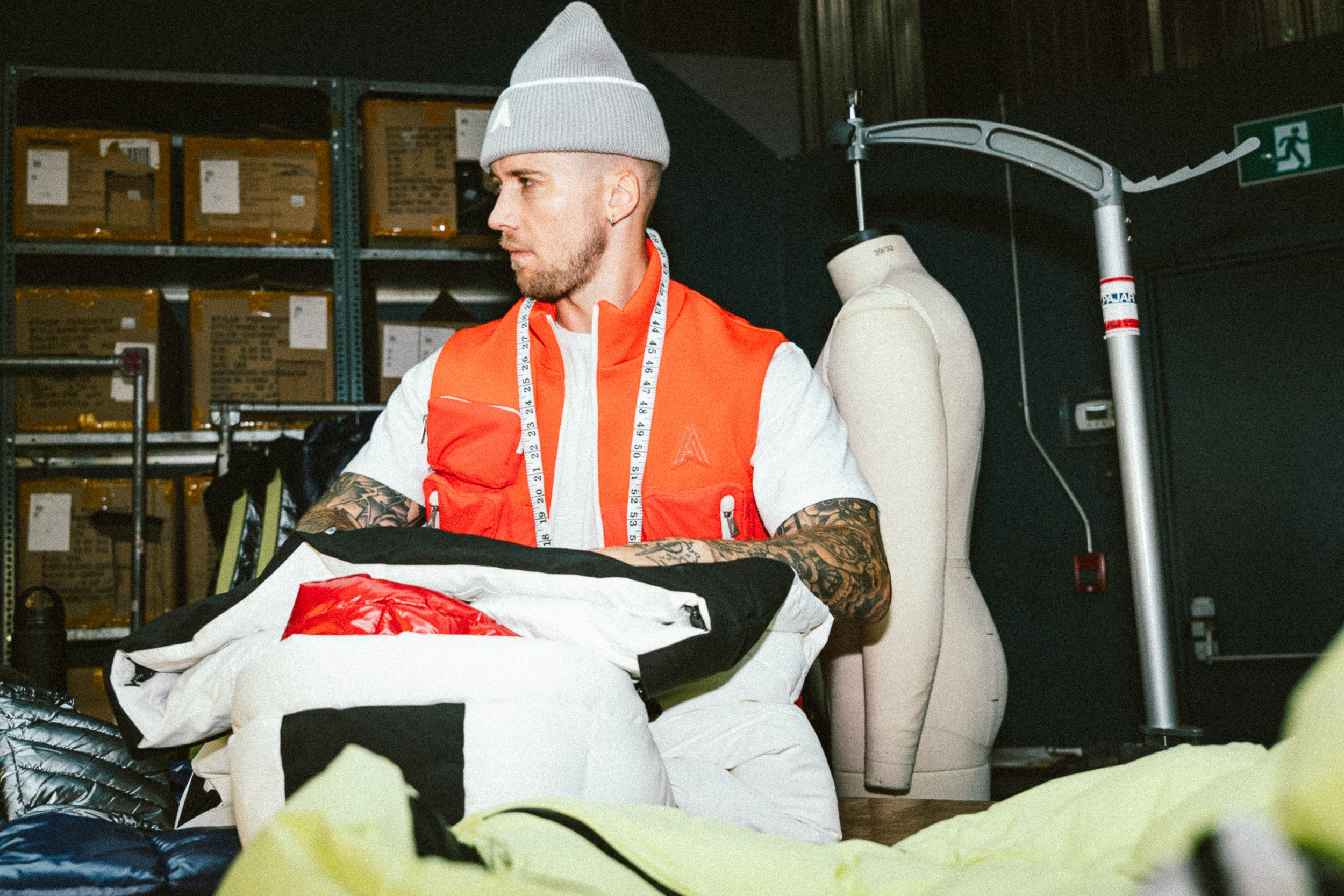 Fashion designer in warehouse, wearing a beanie and red vest, measuring clothes.