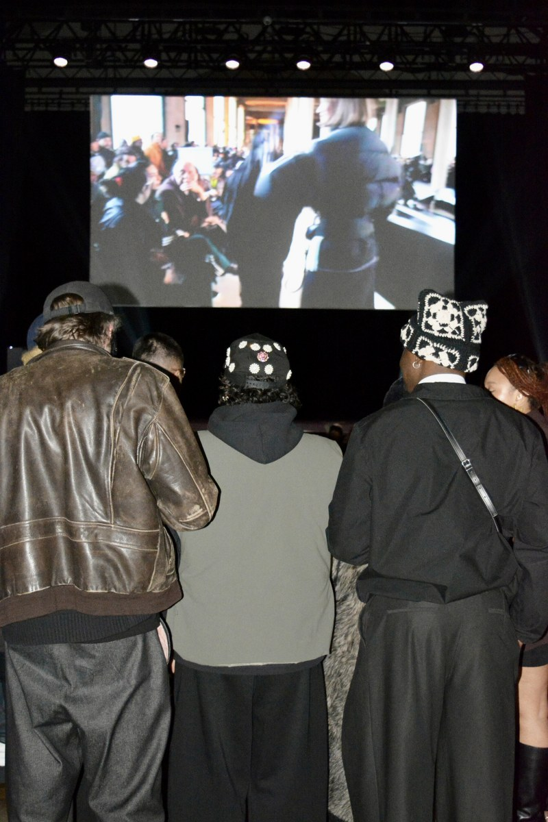 Audience watching a fashion show video at an indoor event with a large screen display.