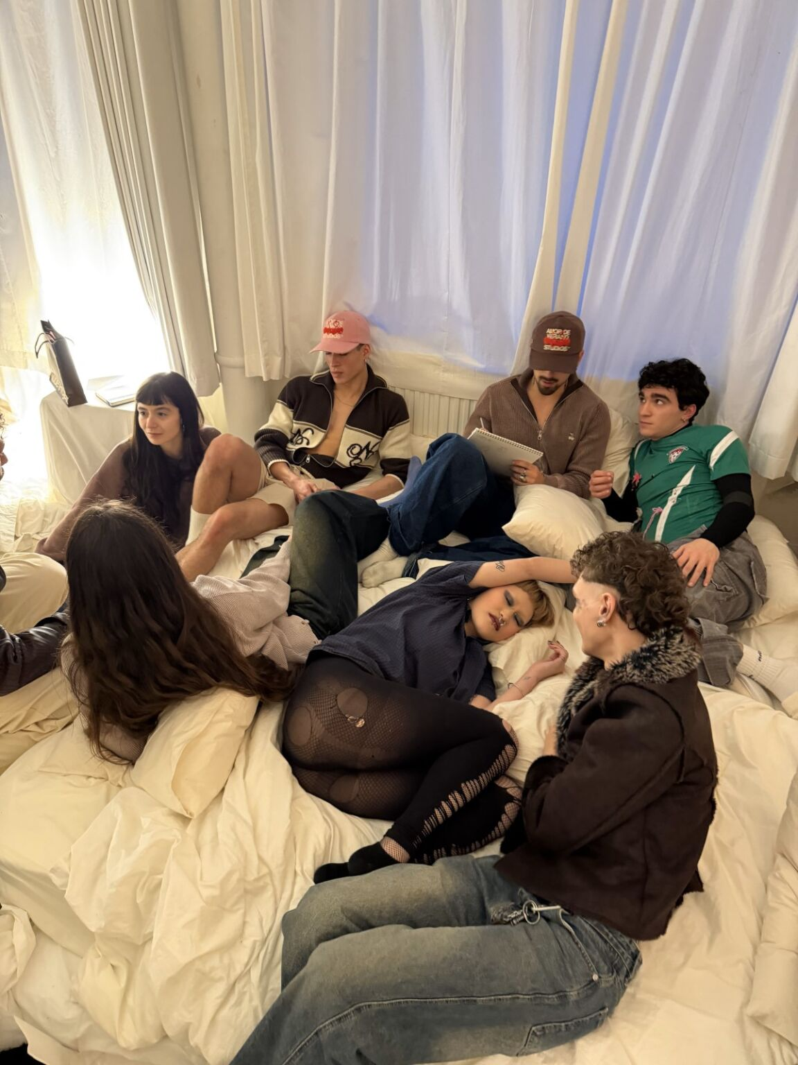 Group of casually dressed friends relaxing on a bed in a cozy room with curtains.