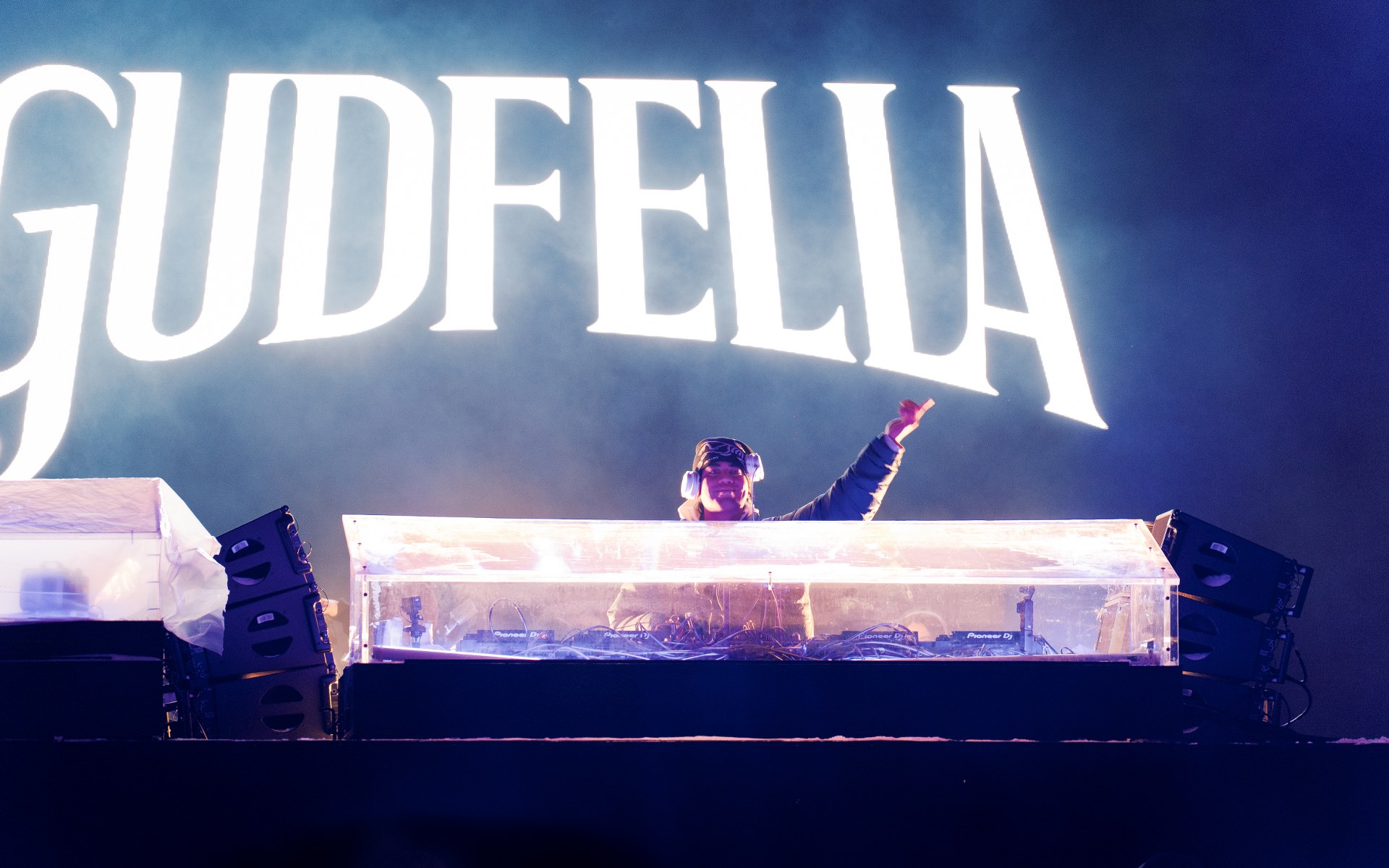 DJ performing on stage with GUDFELLA in the background, vibrant lighting setup.