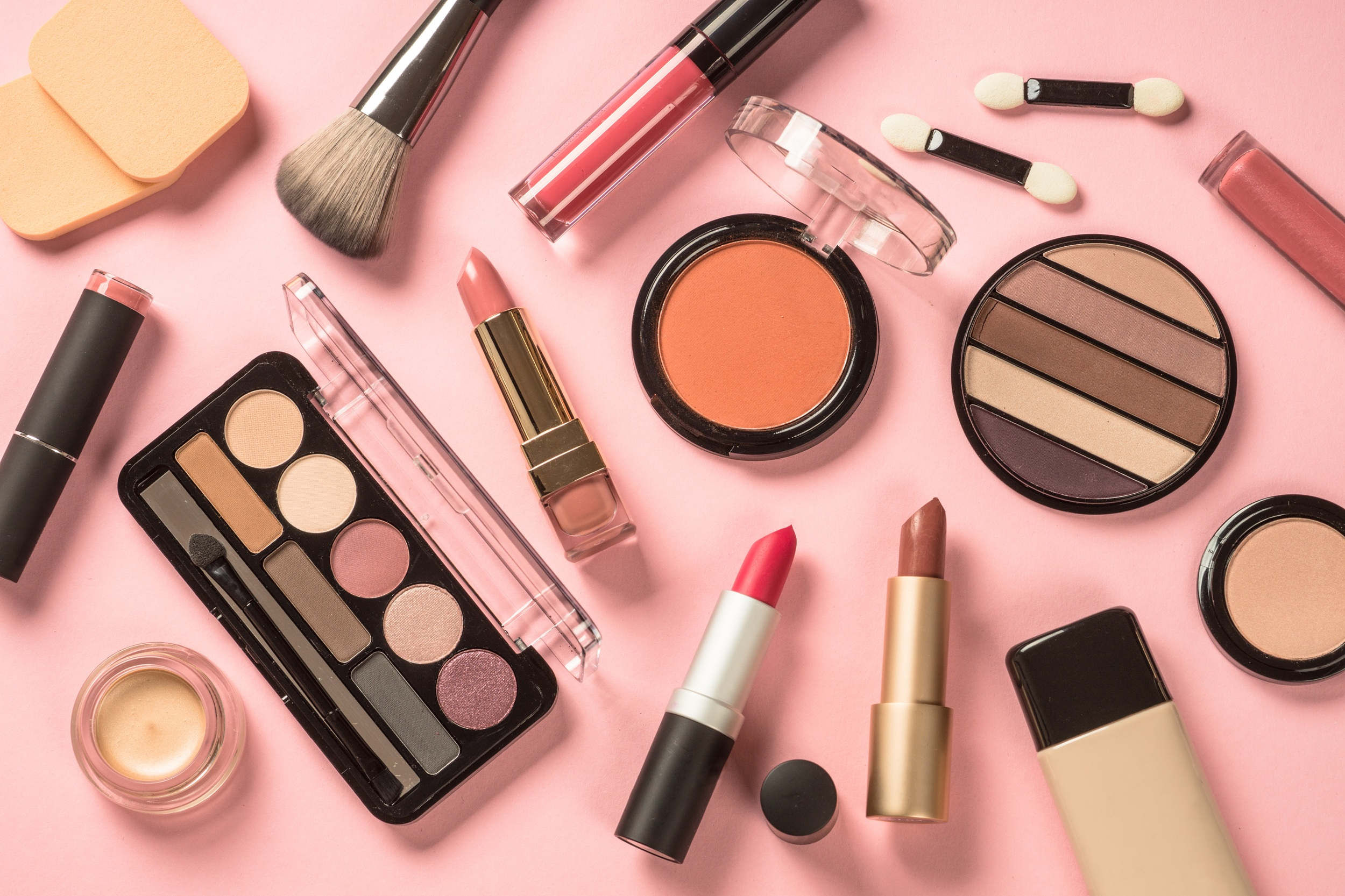 Assorted makeup items on a pink background, including lipsticks and eyeshadow palettes.