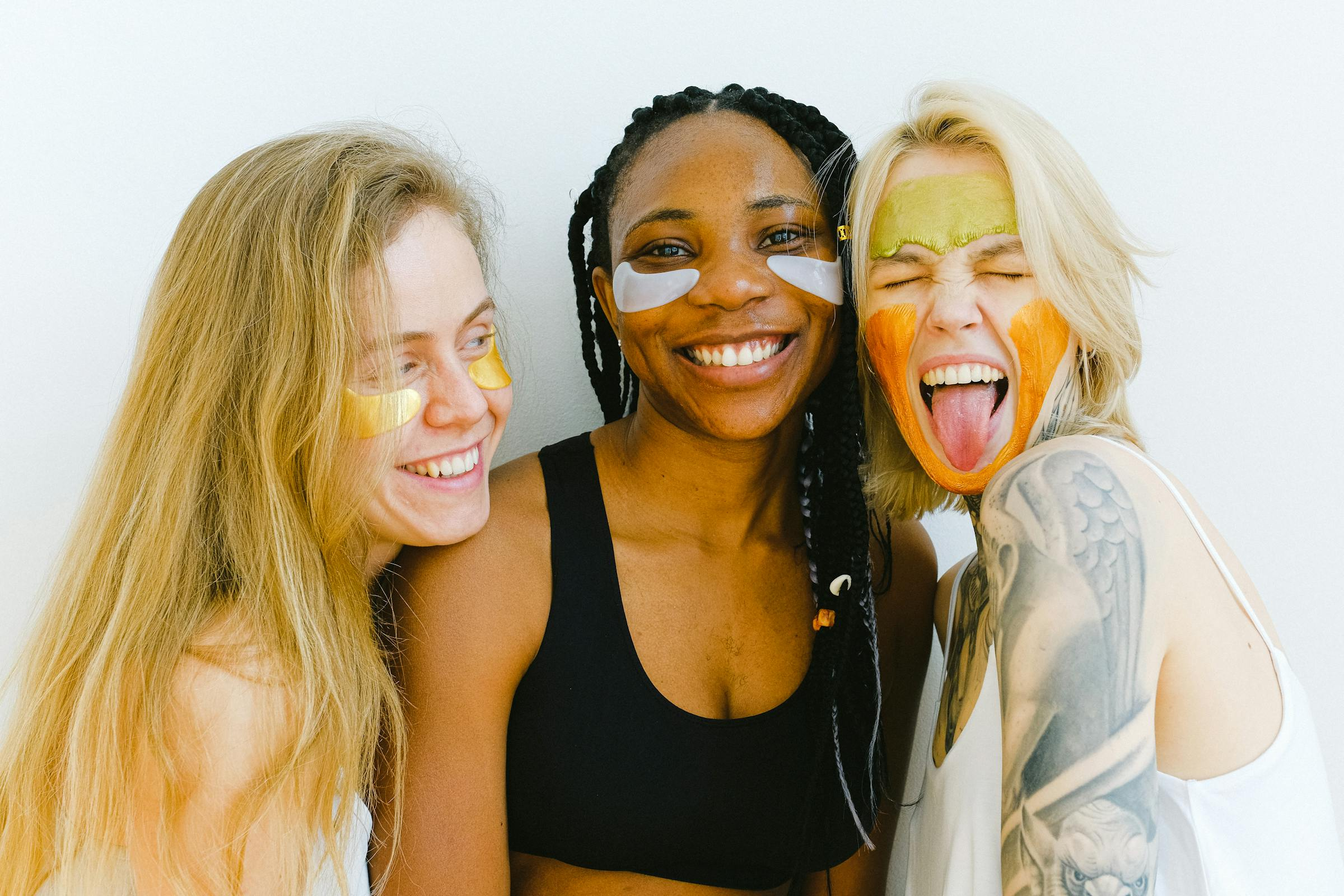 Three women smiling with colorful face masks and under-eye patches.