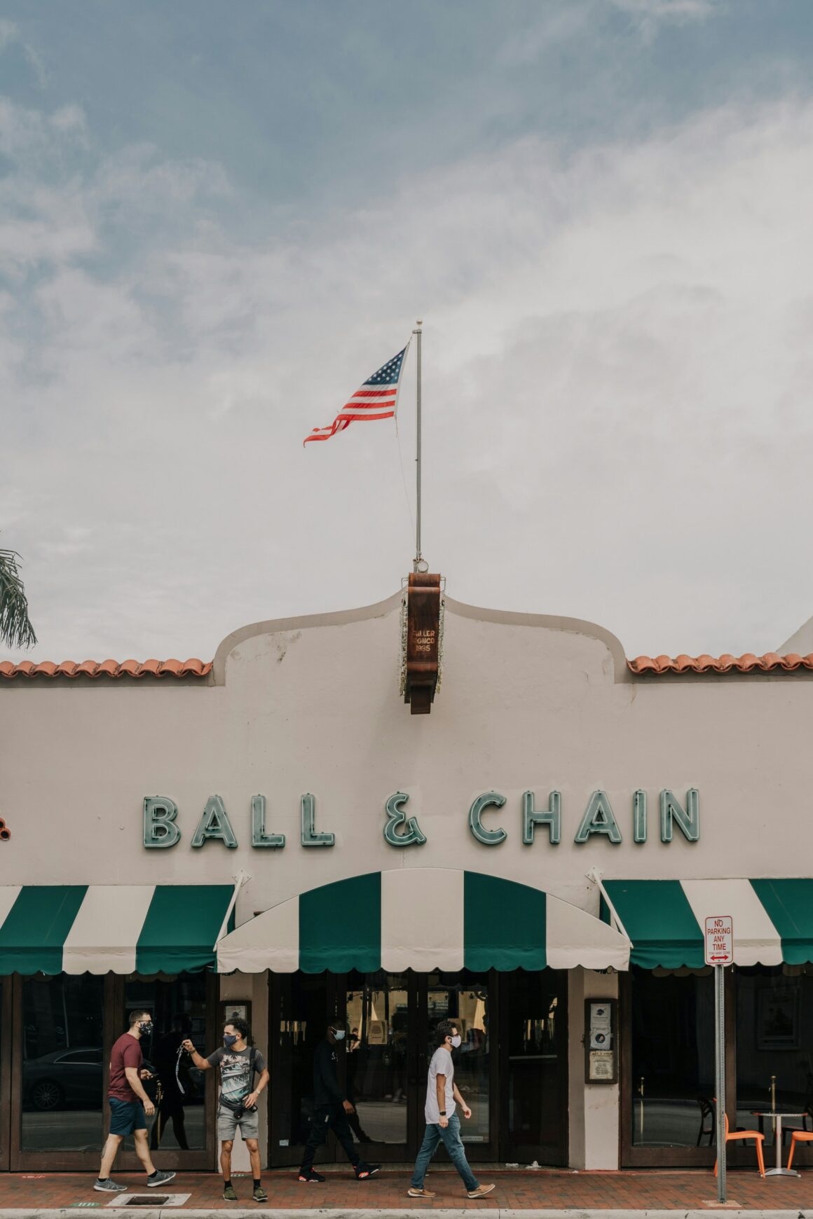 Ball & Chain entrance with American flag, people walking by, blue sky background.