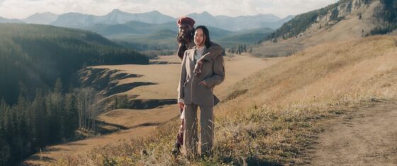 Two people in stylish outfits stand in a picturesque mountain landscape.