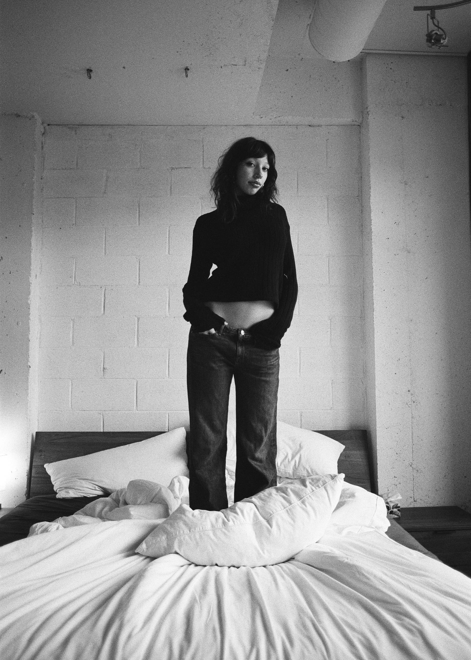 Monochrome photo of a woman standing on a bed, wearing jeans and a black sweater.