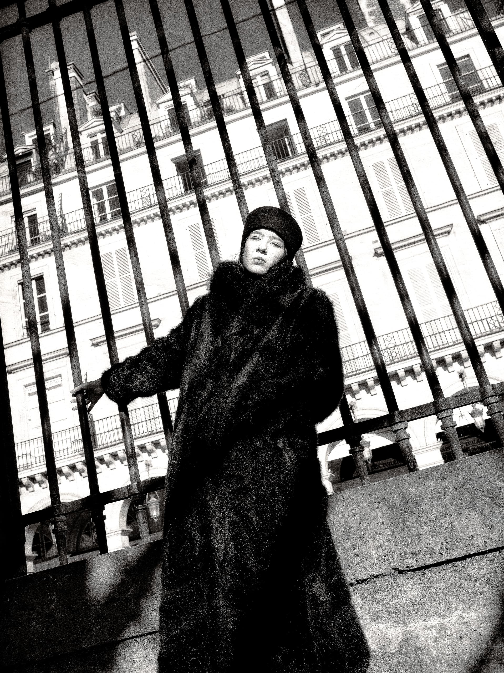 Fashionable person in fur coat standing by wrought iron fence, elegant building behind.
