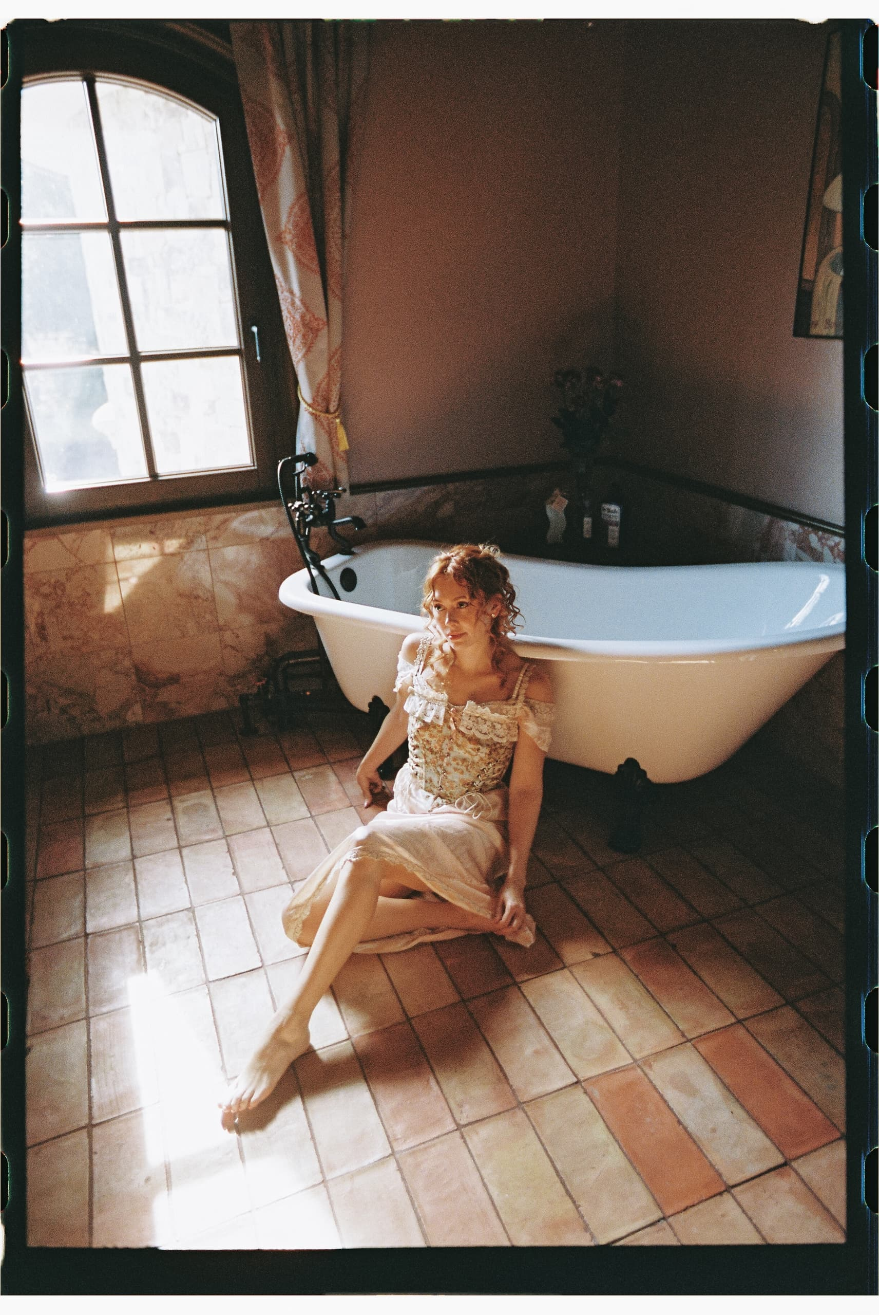 Woman in vintage dress sitting by bathtub in cozy, sunlit bathroom.