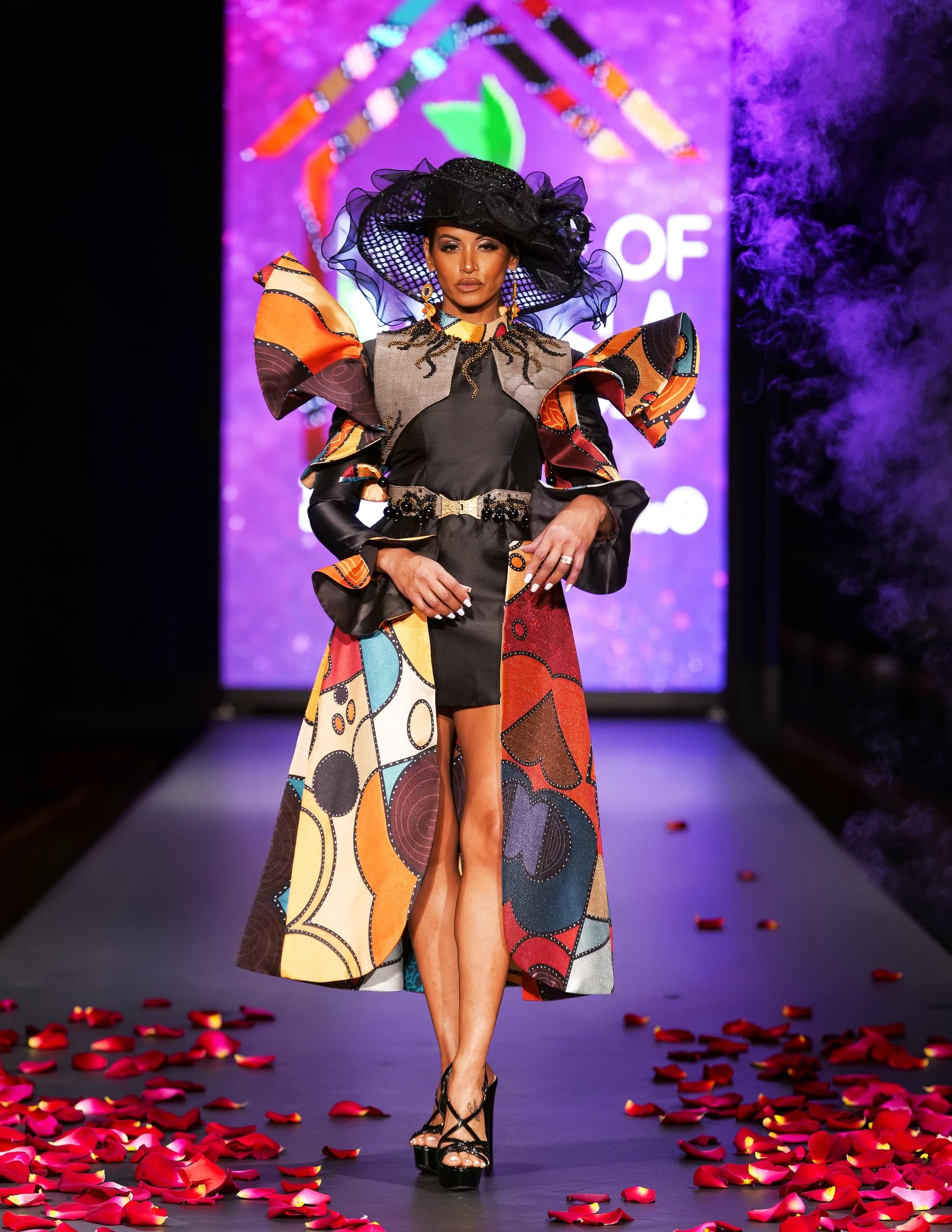 Model in vibrant dress with hat on runway, surrounded by red petals and purple backdrop.
