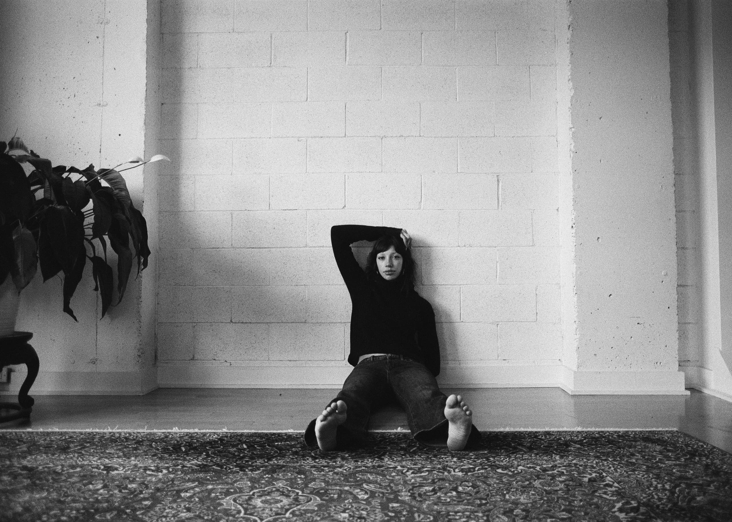 Person sitting against a wall in a minimalist room with a plant. Black and white photography.