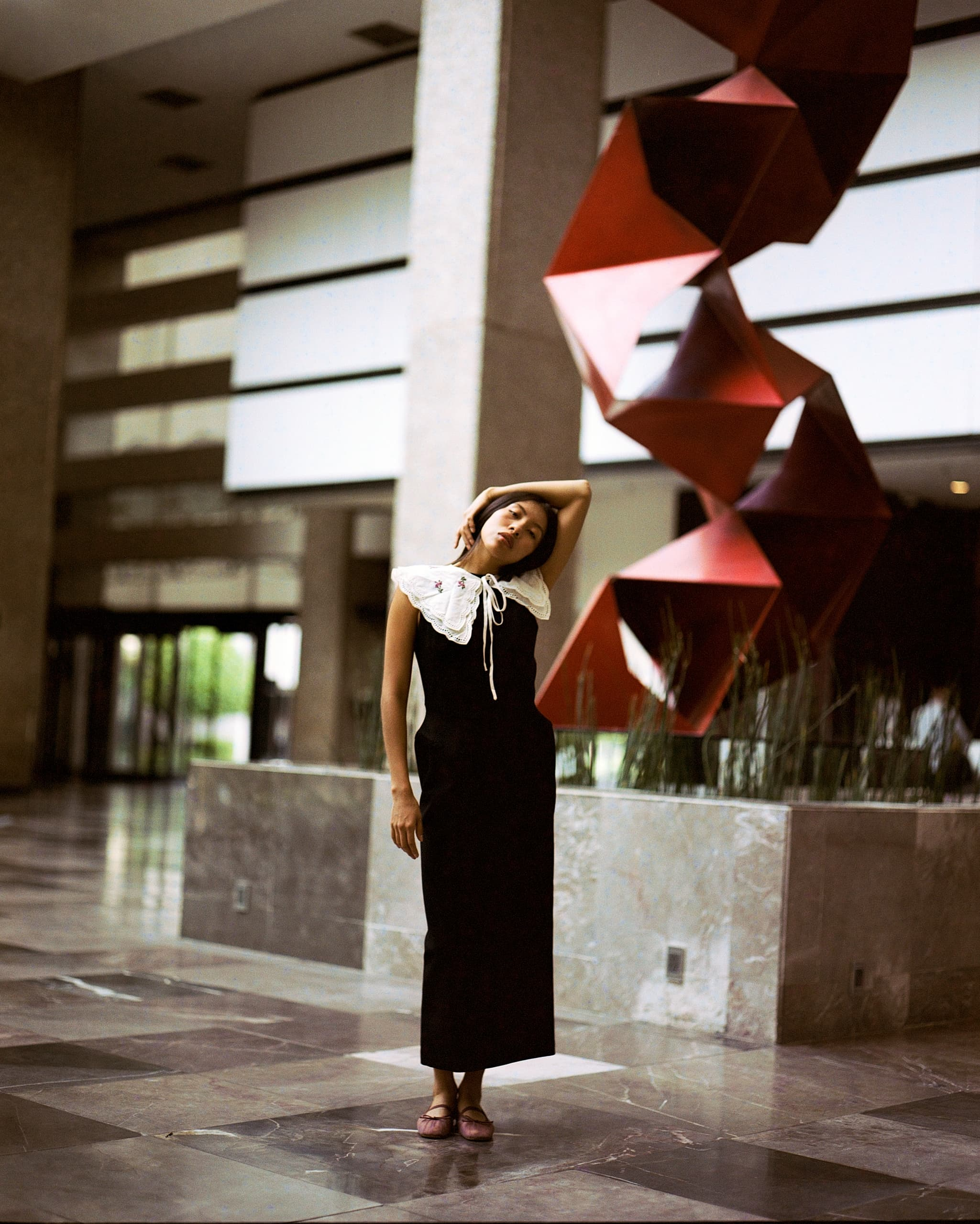 Woman posing in elegant outfit near modern red sculpture in a spacious lobby.