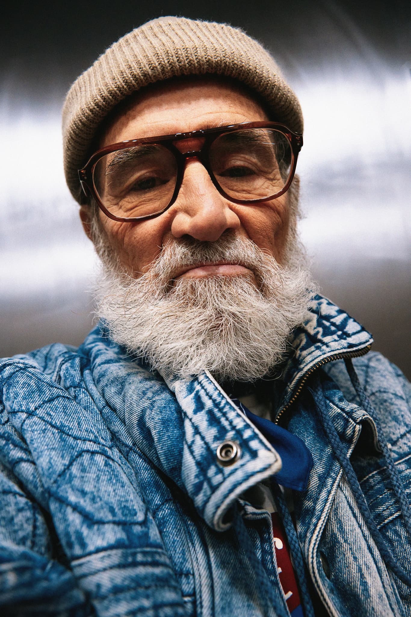 Elderly man in beanie and glasses wearing a blue denim jacket, close-up shot.