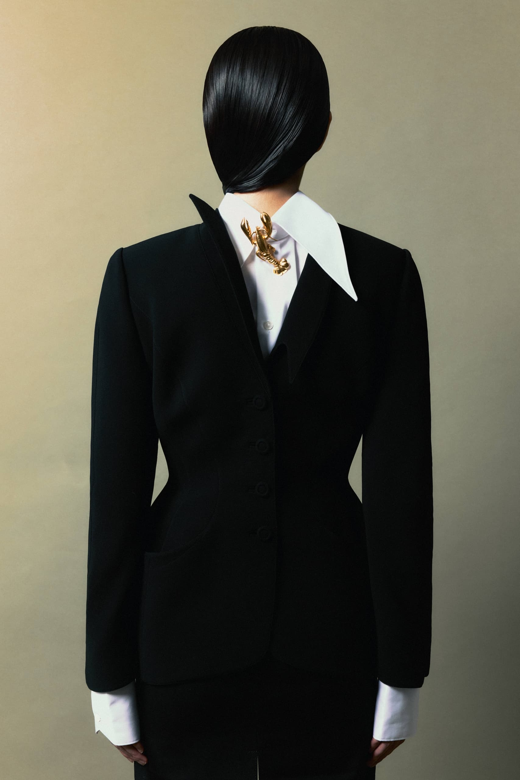 Back view of stylish woman in black suit with gold scorpion brooch on white collar.