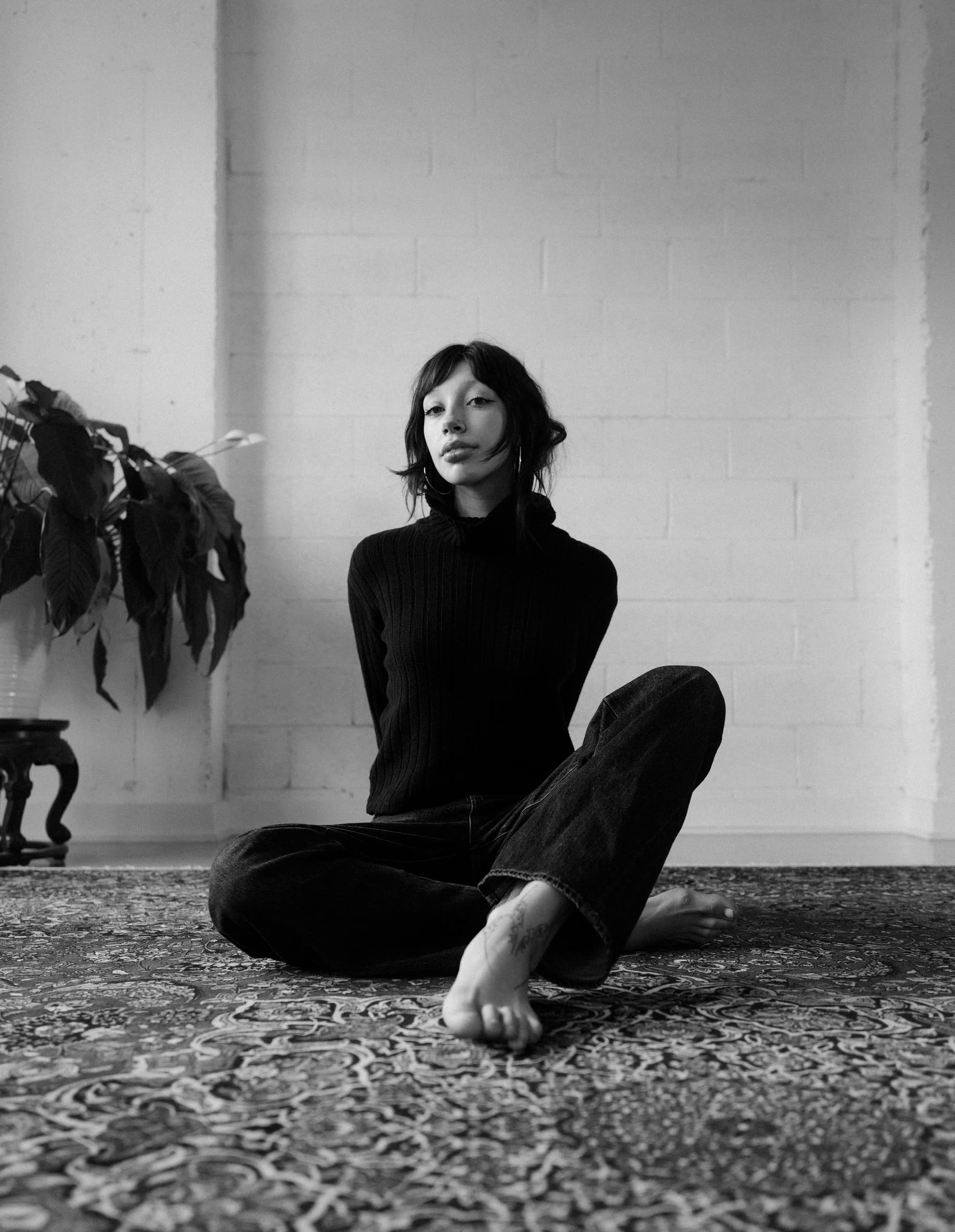 Person in black attire sitting cross-legged on a patterned rug in minimalist space.