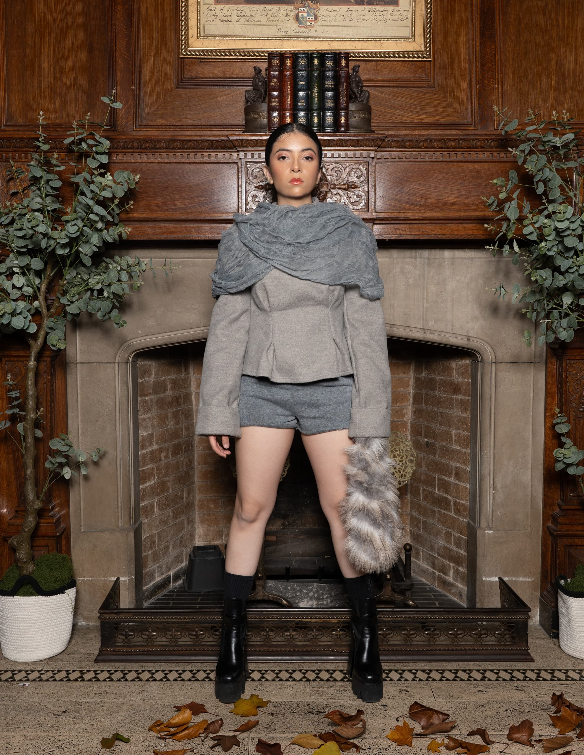 Woman in stylish outfit stands by fireplace with books and plants in a cozy room.