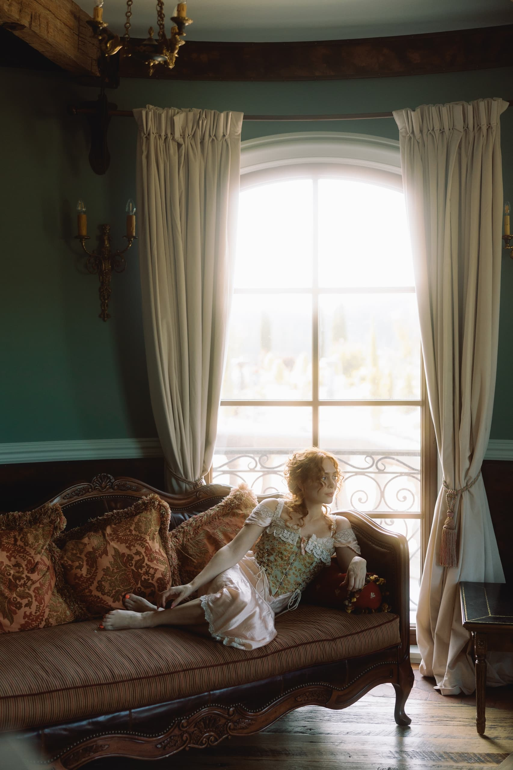 Woman in vintage dress lounging on an antique sofa by a window.