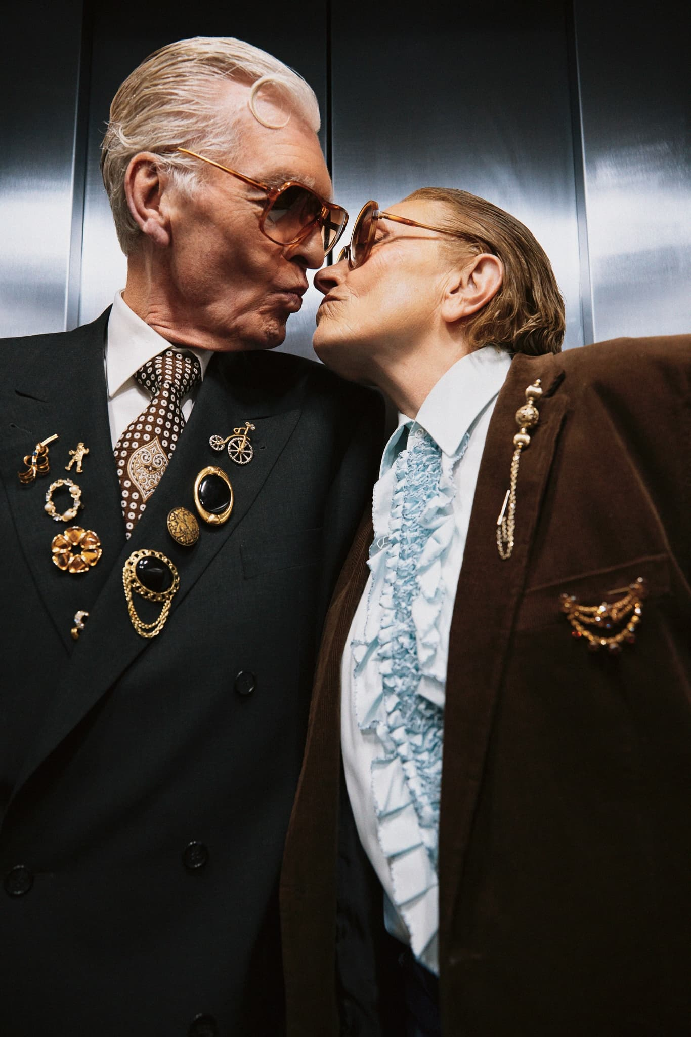 Elderly couple in stylish suits share a tender kiss, adorned with vintage pins.
