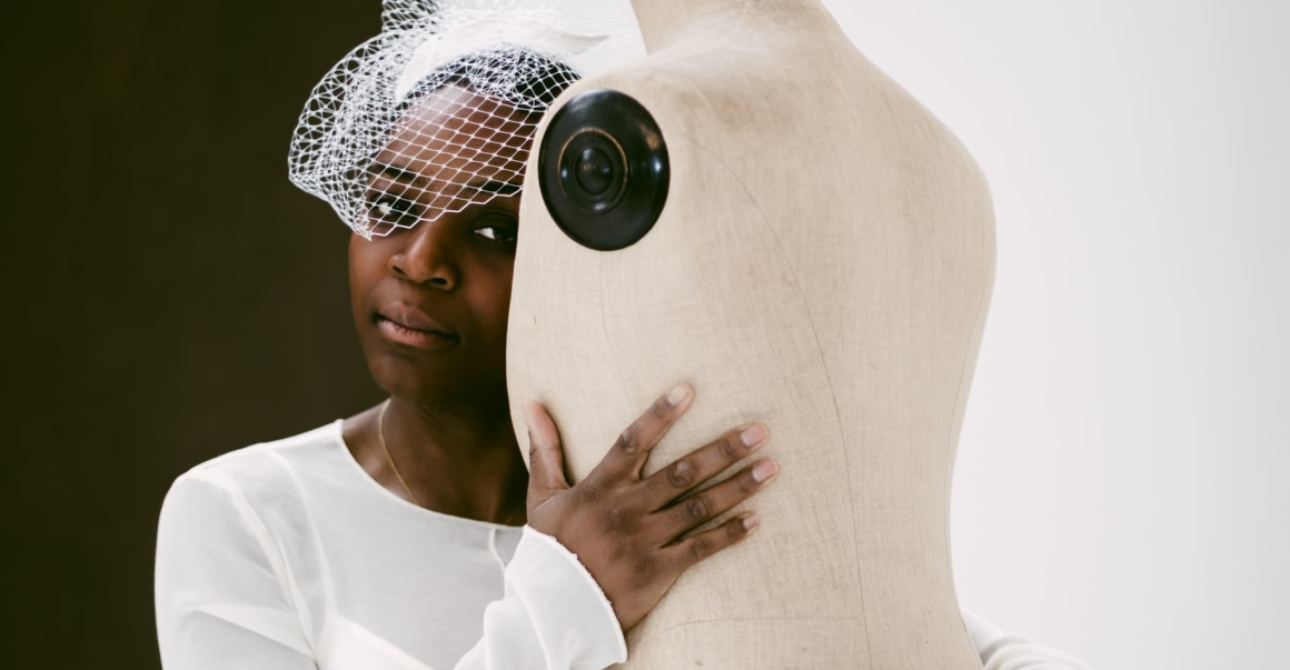 Woman in white dress embraces a mannequin, wearing a netted headpiece.