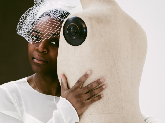 Woman in white dress embraces a mannequin, wearing a netted headpiece.